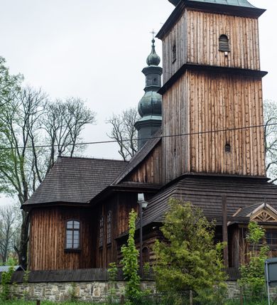 Zdjęcie nr 30: Kościół drewniany o konstrukcji zrębowej, elewacje jednolite, szalowane z listwowaniem, wzmocnione lisicami, wieża o konstrukcji słupowej. Więźba dachowa drewniana, krokwiowa. Budynek na kamiennym podmurowaniu przesłoniętym daszkiem gontowym. Kościół orientowany, jednonawowy z dwiema kaplicami o charakterze transeptu zamkniętymi wielobocznie, na planie krzyża łacińskiego, o węższym prezbiterium zamkniętym trójbocznie. Do wschodniej ściany prezbiterium przylega ogrójec w formie kapliczki, od północy zakrystia na planie prostokąta. Na skrzyżowaniu nawy z południowym ramieniem transeptu kruchta na planie prostokąta. Od zachodu do nawy przylega wieża na planie kwadratu, otoczona sobotami. Prezbiterium i nawa nakryte pozornym sklepieniem tej samej szerokości, obwiedzionym dużym, profilowanym gzymsem podtrzymywanym w nawie przez dwie pary filarów o kanelowanych trzonach, na cokołach. Sklepieniu nawy głównej towarzyszą płaskie i niższe stropy z fasetami w przestrzeniach bocznych. Ściany boczne nawy zwieńczone profilowanym gzymsem. Pomiędzy nawą a prezbiterium otwór tęczowy w formie odcinkowej arkady podtrzymywanej przez filary o kanelowanych trzonach z belką o falistym zarysie. Kaplice boczne otwarte do nawy odcinkowymi arkadami podtrzymywanymi przez filary o kanelowanych trzonach, na cokołach. Parapet chóru muzycznego w zachodniej części nawy, o wybrzuszonej środkowej części, podparty na dwóch filarach o kanelowanych trzonach, na cokołach. Posadzka z płyt ceramicznych. Elewacja frontowa w postaci wieży otoczonej sobotami. Na osi frontowej ściany sobót płytka kruchta zwieńczona gzymsem z przyczółkiem wypełnionym motywem „słoneczka”, przykryta dwuspadowym dachem. Drzwi dwuskrzydłowe, drewniane z okuciami. Korpus wieży podzielony poziomo daszkiem okapowym w dwóch trzecich wysokości, na froncie okno. W dolnej części małe otwory dzwonne z trzech stron, w górnej części duże otwory dzwonne z czterech stron. Wieża zwieńczona hełmem baniastym z latarnią. Ściany sobót szalowane z poziomym listwowaniem, przekryte mansardowym dachem gontowym, na północnej i południowej ścianie dwie pary okien w formie leżącego prostokąta zamkniętego łukiem odcinkowym. W elewacji południowej cztery otwory okienne w formie stojącego prostokąta zamkniętego łukiem odcinkowym, w partii prezbiterium z witrażami. Na elewacji wschodniej ogrójec w formie nadwieszonej kapliczki nakrytej dwuspadowym daszkiem z grupą Ukrzyżowania. W elewacji północnej dwa otwory okienne w formie stojącego prostokąta zamkniętego łukiem odcinkowym. W zakrystii małe kwadratowe okienko. W obu ramionach transeptu analogiczne jak w elewacjach otwory okienne na wschodniej i zachodniej ścianie. Kościół nakryty dachami gontowymi, wyższym nad nawą, niższym nad prezbiterium, dwuspadowym nad nawą oraz wielospadowym nad prezbiterium i ramionami transeptu; nad zakrystią i kruchtą boczną dachy dwuspadowe. W połowie długości dachu, nad nawą, od strony prezbiterium wznosi się ponad kalenicą wieżyczka na sygnaturkę, obita blachą, na ośmiobocznym trzonie, z latarnią zwieńczoną niewielkim baniastym hełmem z latarnią.
