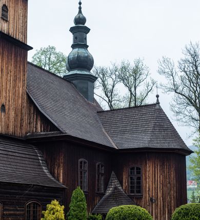 Zdjęcie nr 27: Kościół drewniany o konstrukcji zrębowej, elewacje jednolite, szalowane z listwowaniem, wzmocnione lisicami, wieża o konstrukcji słupowej. Więźba dachowa drewniana, krokwiowa. Budynek na kamiennym podmurowaniu przesłoniętym daszkiem gontowym. Kościół orientowany, jednonawowy z dwiema kaplicami o charakterze transeptu zamkniętymi wielobocznie, na planie krzyża łacińskiego, o węższym prezbiterium zamkniętym trójbocznie. Do wschodniej ściany prezbiterium przylega ogrójec w formie kapliczki, od północy zakrystia na planie prostokąta. Na skrzyżowaniu nawy z południowym ramieniem transeptu kruchta na planie prostokąta. Od zachodu do nawy przylega wieża na planie kwadratu, otoczona sobotami. Prezbiterium i nawa nakryte pozornym sklepieniem tej samej szerokości, obwiedzionym dużym, profilowanym gzymsem podtrzymywanym w nawie przez dwie pary filarów o kanelowanych trzonach, na cokołach. Sklepieniu nawy głównej towarzyszą płaskie i niższe stropy z fasetami w przestrzeniach bocznych. Ściany boczne nawy zwieńczone profilowanym gzymsem. Pomiędzy nawą a prezbiterium otwór tęczowy w formie odcinkowej arkady podtrzymywanej przez filary o kanelowanych trzonach z belką o falistym zarysie. Kaplice boczne otwarte do nawy odcinkowymi arkadami podtrzymywanymi przez filary o kanelowanych trzonach, na cokołach. Parapet chóru muzycznego w zachodniej części nawy, o wybrzuszonej środkowej części, podparty na dwóch filarach o kanelowanych trzonach, na cokołach. Posadzka z płyt ceramicznych. Elewacja frontowa w postaci wieży otoczonej sobotami. Na osi frontowej ściany sobót płytka kruchta zwieńczona gzymsem z przyczółkiem wypełnionym motywem „słoneczka”, przykryta dwuspadowym dachem. Drzwi dwuskrzydłowe, drewniane z okuciami. Korpus wieży podzielony poziomo daszkiem okapowym w dwóch trzecich wysokości, na froncie okno. W dolnej części małe otwory dzwonne z trzech stron, w górnej części duże otwory dzwonne z czterech stron. Wieża zwieńczona hełmem baniastym z latarnią. Ściany sobót szalowane z poziomym listwowaniem, przekryte mansardowym dachem gontowym, na północnej i południowej ścianie dwie pary okien w formie leżącego prostokąta zamkniętego łukiem odcinkowym. W elewacji południowej cztery otwory okienne w formie stojącego prostokąta zamkniętego łukiem odcinkowym, w partii prezbiterium z witrażami. Na elewacji wschodniej ogrójec w formie nadwieszonej kapliczki nakrytej dwuspadowym daszkiem z grupą Ukrzyżowania. W elewacji północnej dwa otwory okienne w formie stojącego prostokąta zamkniętego łukiem odcinkowym. W zakrystii małe kwadratowe okienko. W obu ramionach transeptu analogiczne jak w elewacjach otwory okienne na wschodniej i zachodniej ścianie. Kościół nakryty dachami gontowymi, wyższym nad nawą, niższym nad prezbiterium, dwuspadowym nad nawą oraz wielospadowym nad prezbiterium i ramionami transeptu; nad zakrystią i kruchtą boczną dachy dwuspadowe. W połowie długości dachu, nad nawą, od strony prezbiterium wznosi się ponad kalenicą wieżyczka na sygnaturkę, obita blachą, na ośmiobocznym trzonie, z latarnią zwieńczoną niewielkim baniastym hełmem z latarnią.
