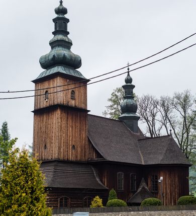 Zdjęcie nr 25: Kościół drewniany o konstrukcji zrębowej, elewacje jednolite, szalowane z listwowaniem, wzmocnione lisicami, wieża o konstrukcji słupowej. Więźba dachowa drewniana, krokwiowa. Budynek na kamiennym podmurowaniu przesłoniętym daszkiem gontowym. Kościół orientowany, jednonawowy z dwiema kaplicami o charakterze transeptu zamkniętymi wielobocznie, na planie krzyża łacińskiego, o węższym prezbiterium zamkniętym trójbocznie. Do wschodniej ściany prezbiterium przylega ogrójec w formie kapliczki, od północy zakrystia na planie prostokąta. Na skrzyżowaniu nawy z południowym ramieniem transeptu kruchta na planie prostokąta. Od zachodu do nawy przylega wieża na planie kwadratu, otoczona sobotami. Prezbiterium i nawa nakryte pozornym sklepieniem tej samej szerokości, obwiedzionym dużym, profilowanym gzymsem podtrzymywanym w nawie przez dwie pary filarów o kanelowanych trzonach, na cokołach. Sklepieniu nawy głównej towarzyszą płaskie i niższe stropy z fasetami w przestrzeniach bocznych. Ściany boczne nawy zwieńczone profilowanym gzymsem. Pomiędzy nawą a prezbiterium otwór tęczowy w formie odcinkowej arkady podtrzymywanej przez filary o kanelowanych trzonach z belką o falistym zarysie. Kaplice boczne otwarte do nawy odcinkowymi arkadami podtrzymywanymi przez filary o kanelowanych trzonach, na cokołach. Parapet chóru muzycznego w zachodniej części nawy, o wybrzuszonej środkowej części, podparty na dwóch filarach o kanelowanych trzonach, na cokołach. Posadzka z płyt ceramicznych. Elewacja frontowa w postaci wieży otoczonej sobotami. Na osi frontowej ściany sobót płytka kruchta zwieńczona gzymsem z przyczółkiem wypełnionym motywem „słoneczka”, przykryta dwuspadowym dachem. Drzwi dwuskrzydłowe, drewniane z okuciami. Korpus wieży podzielony poziomo daszkiem okapowym w dwóch trzecich wysokości, na froncie okno. W dolnej części małe otwory dzwonne z trzech stron, w górnej części duże otwory dzwonne z czterech stron. Wieża zwieńczona hełmem baniastym z latarnią. Ściany sobót szalowane z poziomym listwowaniem, przekryte mansardowym dachem gontowym, na północnej i południowej ścianie dwie pary okien w formie leżącego prostokąta zamkniętego łukiem odcinkowym. W elewacji południowej cztery otwory okienne w formie stojącego prostokąta zamkniętego łukiem odcinkowym, w partii prezbiterium z witrażami. Na elewacji wschodniej ogrójec w formie nadwieszonej kapliczki nakrytej dwuspadowym daszkiem z grupą Ukrzyżowania. W elewacji północnej dwa otwory okienne w formie stojącego prostokąta zamkniętego łukiem odcinkowym. W zakrystii małe kwadratowe okienko. W obu ramionach transeptu analogiczne jak w elewacjach otwory okienne na wschodniej i zachodniej ścianie. Kościół nakryty dachami gontowymi, wyższym nad nawą, niższym nad prezbiterium, dwuspadowym nad nawą oraz wielospadowym nad prezbiterium i ramionami transeptu; nad zakrystią i kruchtą boczną dachy dwuspadowe. W połowie długości dachu, nad nawą, od strony prezbiterium wznosi się ponad kalenicą wieżyczka na sygnaturkę, obita blachą, na ośmiobocznym trzonie, z latarnią zwieńczoną niewielkim baniastym hełmem z latarnią.

