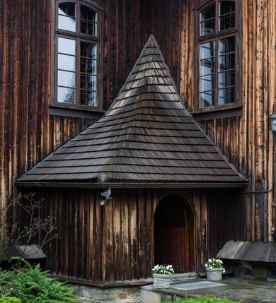 Zdjęcie nr 50: Kościół drewniany o konstrukcji zrębowej, elewacje jednolite, szalowane z listwowaniem, wzmocnione lisicami, wieża o konstrukcji słupowej. Więźba dachowa drewniana, krokwiowa. Budynek na kamiennym podmurowaniu przesłoniętym daszkiem gontowym. Kościół orientowany, jednonawowy z dwiema kaplicami o charakterze transeptu zamkniętymi wielobocznie, na planie krzyża łacińskiego, o węższym prezbiterium zamkniętym trójbocznie. Do wschodniej ściany prezbiterium przylega ogrójec w formie kapliczki, od północy zakrystia na planie prostokąta. Na skrzyżowaniu nawy z południowym ramieniem transeptu kruchta na planie prostokąta. Od zachodu do nawy przylega wieża na planie kwadratu, otoczona sobotami. Prezbiterium i nawa nakryte pozornym sklepieniem tej samej szerokości, obwiedzionym dużym, profilowanym gzymsem podtrzymywanym w nawie przez dwie pary filarów o kanelowanych trzonach, na cokołach. Sklepieniu nawy głównej towarzyszą płaskie i niższe stropy z fasetami w przestrzeniach bocznych. Ściany boczne nawy zwieńczone profilowanym gzymsem. Pomiędzy nawą a prezbiterium otwór tęczowy w formie odcinkowej arkady podtrzymywanej przez filary o kanelowanych trzonach z belką o falistym zarysie. Kaplice boczne otwarte do nawy odcinkowymi arkadami podtrzymywanymi przez filary o kanelowanych trzonach, na cokołach. Parapet chóru muzycznego w zachodniej części nawy, o wybrzuszonej środkowej części, podparty na dwóch filarach o kanelowanych trzonach, na cokołach. Posadzka z płyt ceramicznych. Elewacja frontowa w postaci wieży otoczonej sobotami. Na osi frontowej ściany sobót płytka kruchta zwieńczona gzymsem z przyczółkiem wypełnionym motywem „słoneczka”, przykryta dwuspadowym dachem. Drzwi dwuskrzydłowe, drewniane z okuciami. Korpus wieży podzielony poziomo daszkiem okapowym w dwóch trzecich wysokości, na froncie okno. W dolnej części małe otwory dzwonne z trzech stron, w górnej części duże otwory dzwonne z czterech stron. Wieża zwieńczona hełmem baniastym z latarnią. Ściany sobót szalowane z poziomym listwowaniem, przekryte mansardowym dachem gontowym, na północnej i południowej ścianie dwie pary okien w formie leżącego prostokąta zamkniętego łukiem odcinkowym. W elewacji południowej cztery otwory okienne w formie stojącego prostokąta zamkniętego łukiem odcinkowym, w partii prezbiterium z witrażami. Na elewacji wschodniej ogrójec w formie nadwieszonej kapliczki nakrytej dwuspadowym daszkiem z grupą Ukrzyżowania. W elewacji północnej dwa otwory okienne w formie stojącego prostokąta zamkniętego łukiem odcinkowym. W zakrystii małe kwadratowe okienko. W obu ramionach transeptu analogiczne jak w elewacjach otwory okienne na wschodniej i zachodniej ścianie. Kościół nakryty dachami gontowymi, wyższym nad nawą, niższym nad prezbiterium, dwuspadowym nad nawą oraz wielospadowym nad prezbiterium i ramionami transeptu; nad zakrystią i kruchtą boczną dachy dwuspadowe. W połowie długości dachu, nad nawą, od strony prezbiterium wznosi się ponad kalenicą wieżyczka na sygnaturkę, obita blachą, na ośmiobocznym trzonie, z latarnią zwieńczoną niewielkim baniastym hełmem z latarnią.
