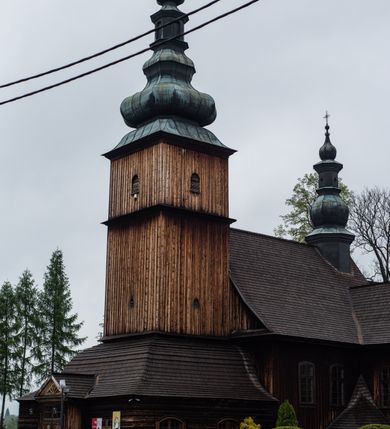 Zdjęcie nr 26: Kościół drewniany o konstrukcji zrębowej, elewacje jednolite, szalowane z listwowaniem, wzmocnione lisicami, wieża o konstrukcji słupowej. Więźba dachowa drewniana, krokwiowa. Budynek na kamiennym podmurowaniu przesłoniętym daszkiem gontowym. Kościół orientowany, jednonawowy z dwiema kaplicami o charakterze transeptu zamkniętymi wielobocznie, na planie krzyża łacińskiego, o węższym prezbiterium zamkniętym trójbocznie. Do wschodniej ściany prezbiterium przylega ogrójec w formie kapliczki, od północy zakrystia na planie prostokąta. Na skrzyżowaniu nawy z południowym ramieniem transeptu kruchta na planie prostokąta. Od zachodu do nawy przylega wieża na planie kwadratu, otoczona sobotami. Prezbiterium i nawa nakryte pozornym sklepieniem tej samej szerokości, obwiedzionym dużym, profilowanym gzymsem podtrzymywanym w nawie przez dwie pary filarów o kanelowanych trzonach, na cokołach. Sklepieniu nawy głównej towarzyszą płaskie i niższe stropy z fasetami w przestrzeniach bocznych. Ściany boczne nawy zwieńczone profilowanym gzymsem. Pomiędzy nawą a prezbiterium otwór tęczowy w formie odcinkowej arkady podtrzymywanej przez filary o kanelowanych trzonach z belką o falistym zarysie. Kaplice boczne otwarte do nawy odcinkowymi arkadami podtrzymywanymi przez filary o kanelowanych trzonach, na cokołach. Parapet chóru muzycznego w zachodniej części nawy, o wybrzuszonej środkowej części, podparty na dwóch filarach o kanelowanych trzonach, na cokołach. Posadzka z płyt ceramicznych. Elewacja frontowa w postaci wieży otoczonej sobotami. Na osi frontowej ściany sobót płytka kruchta zwieńczona gzymsem z przyczółkiem wypełnionym motywem „słoneczka”, przykryta dwuspadowym dachem. Drzwi dwuskrzydłowe, drewniane z okuciami. Korpus wieży podzielony poziomo daszkiem okapowym w dwóch trzecich wysokości, na froncie okno. W dolnej części małe otwory dzwonne z trzech stron, w górnej części duże otwory dzwonne z czterech stron. Wieża zwieńczona hełmem baniastym z latarnią. Ściany sobót szalowane z poziomym listwowaniem, przekryte mansardowym dachem gontowym, na północnej i południowej ścianie dwie pary okien w formie leżącego prostokąta zamkniętego łukiem odcinkowym. W elewacji południowej cztery otwory okienne w formie stojącego prostokąta zamkniętego łukiem odcinkowym, w partii prezbiterium z witrażami. Na elewacji wschodniej ogrójec w formie nadwieszonej kapliczki nakrytej dwuspadowym daszkiem z grupą Ukrzyżowania. W elewacji północnej dwa otwory okienne w formie stojącego prostokąta zamkniętego łukiem odcinkowym. W zakrystii małe kwadratowe okienko. W obu ramionach transeptu analogiczne jak w elewacjach otwory okienne na wschodniej i zachodniej ścianie. Kościół nakryty dachami gontowymi, wyższym nad nawą, niższym nad prezbiterium, dwuspadowym nad nawą oraz wielospadowym nad prezbiterium i ramionami transeptu; nad zakrystią i kruchtą boczną dachy dwuspadowe. W połowie długości dachu, nad nawą, od strony prezbiterium wznosi się ponad kalenicą wieżyczka na sygnaturkę, obita blachą, na ośmiobocznym trzonie, z latarnią zwieńczoną niewielkim baniastym hełmem z latarnią.
