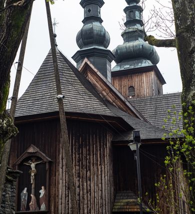 Zdjęcie nr 51: Kościół drewniany o konstrukcji zrębowej, elewacje jednolite, szalowane z listwowaniem, wzmocnione lisicami, wieża o konstrukcji słupowej. Więźba dachowa drewniana, krokwiowa. Budynek na kamiennym podmurowaniu przesłoniętym daszkiem gontowym. Kościół orientowany, jednonawowy z dwiema kaplicami o charakterze transeptu zamkniętymi wielobocznie, na planie krzyża łacińskiego, o węższym prezbiterium zamkniętym trójbocznie. Do wschodniej ściany prezbiterium przylega ogrójec w formie kapliczki, od północy zakrystia na planie prostokąta. Na skrzyżowaniu nawy z południowym ramieniem transeptu kruchta na planie prostokąta. Od zachodu do nawy przylega wieża na planie kwadratu, otoczona sobotami. Prezbiterium i nawa nakryte pozornym sklepieniem tej samej szerokości, obwiedzionym dużym, profilowanym gzymsem podtrzymywanym w nawie przez dwie pary filarów o kanelowanych trzonach, na cokołach. Sklepieniu nawy głównej towarzyszą płaskie i niższe stropy z fasetami w przestrzeniach bocznych. Ściany boczne nawy zwieńczone profilowanym gzymsem. Pomiędzy nawą a prezbiterium otwór tęczowy w formie odcinkowej arkady podtrzymywanej przez filary o kanelowanych trzonach z belką o falistym zarysie. Kaplice boczne otwarte do nawy odcinkowymi arkadami podtrzymywanymi przez filary o kanelowanych trzonach, na cokołach. Parapet chóru muzycznego w zachodniej części nawy, o wybrzuszonej środkowej części, podparty na dwóch filarach o kanelowanych trzonach, na cokołach. Posadzka z płyt ceramicznych. Elewacja frontowa w postaci wieży otoczonej sobotami. Na osi frontowej ściany sobót płytka kruchta zwieńczona gzymsem z przyczółkiem wypełnionym motywem „słoneczka”, przykryta dwuspadowym dachem. Drzwi dwuskrzydłowe, drewniane z okuciami. Korpus wieży podzielony poziomo daszkiem okapowym w dwóch trzecich wysokości, na froncie okno. W dolnej części małe otwory dzwonne z trzech stron, w górnej części duże otwory dzwonne z czterech stron. Wieża zwieńczona hełmem baniastym z latarnią. Ściany sobót szalowane z poziomym listwowaniem, przekryte mansardowym dachem gontowym, na północnej i południowej ścianie dwie pary okien w formie leżącego prostokąta zamkniętego łukiem odcinkowym. W elewacji południowej cztery otwory okienne w formie stojącego prostokąta zamkniętego łukiem odcinkowym, w partii prezbiterium z witrażami. Na elewacji wschodniej ogrójec w formie nadwieszonej kapliczki nakrytej dwuspadowym daszkiem z grupą Ukrzyżowania. W elewacji północnej dwa otwory okienne w formie stojącego prostokąta zamkniętego łukiem odcinkowym. W zakrystii małe kwadratowe okienko. W obu ramionach transeptu analogiczne jak w elewacjach otwory okienne na wschodniej i zachodniej ścianie. Kościół nakryty dachami gontowymi, wyższym nad nawą, niższym nad prezbiterium, dwuspadowym nad nawą oraz wielospadowym nad prezbiterium i ramionami transeptu; nad zakrystią i kruchtą boczną dachy dwuspadowe. W połowie długości dachu, nad nawą, od strony prezbiterium wznosi się ponad kalenicą wieżyczka na sygnaturkę, obita blachą, na ośmiobocznym trzonie, z latarnią zwieńczoną niewielkim baniastym hełmem z latarnią.
