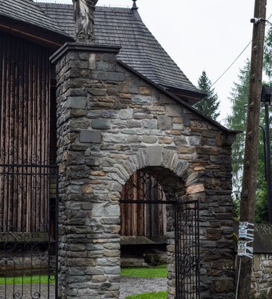 Zdjęcie nr 58: Kościół drewniany o konstrukcji zrębowej, elewacje jednolite, szalowane z listwowaniem, wzmocnione lisicami, wieża o konstrukcji słupowej. Więźba dachowa drewniana, krokwiowa. Budynek na kamiennym podmurowaniu przesłoniętym daszkiem gontowym. Kościół orientowany, jednonawowy z dwiema kaplicami o charakterze transeptu zamkniętymi wielobocznie, na planie krzyża łacińskiego, o węższym prezbiterium zamkniętym trójbocznie. Do wschodniej ściany prezbiterium przylega ogrójec w formie kapliczki, od północy zakrystia na planie prostokąta. Na skrzyżowaniu nawy z południowym ramieniem transeptu kruchta na planie prostokąta. Od zachodu do nawy przylega wieża na planie kwadratu, otoczona sobotami. Prezbiterium i nawa nakryte pozornym sklepieniem tej samej szerokości, obwiedzionym dużym, profilowanym gzymsem podtrzymywanym w nawie przez dwie pary filarów o kanelowanych trzonach, na cokołach. Sklepieniu nawy głównej towarzyszą płaskie i niższe stropy z fasetami w przestrzeniach bocznych. Ściany boczne nawy zwieńczone profilowanym gzymsem. Pomiędzy nawą a prezbiterium otwór tęczowy w formie odcinkowej arkady podtrzymywanej przez filary o kanelowanych trzonach z belką o falistym zarysie. Kaplice boczne otwarte do nawy odcinkowymi arkadami podtrzymywanymi przez filary o kanelowanych trzonach, na cokołach. Parapet chóru muzycznego w zachodniej części nawy, o wybrzuszonej środkowej części, podparty na dwóch filarach o kanelowanych trzonach, na cokołach. Posadzka z płyt ceramicznych. Elewacja frontowa w postaci wieży otoczonej sobotami. Na osi frontowej ściany sobót płytka kruchta zwieńczona gzymsem z przyczółkiem wypełnionym motywem „słoneczka”, przykryta dwuspadowym dachem. Drzwi dwuskrzydłowe, drewniane z okuciami. Korpus wieży podzielony poziomo daszkiem okapowym w dwóch trzecich wysokości, na froncie okno. W dolnej części małe otwory dzwonne z trzech stron, w górnej części duże otwory dzwonne z czterech stron. Wieża zwieńczona hełmem baniastym z latarnią. Ściany sobót szalowane z poziomym listwowaniem, przekryte mansardowym dachem gontowym, na północnej i południowej ścianie dwie pary okien w formie leżącego prostokąta zamkniętego łukiem odcinkowym. W elewacji południowej cztery otwory okienne w formie stojącego prostokąta zamkniętego łukiem odcinkowym, w partii prezbiterium z witrażami. Na elewacji wschodniej ogrójec w formie nadwieszonej kapliczki nakrytej dwuspadowym daszkiem z grupą Ukrzyżowania. W elewacji północnej dwa otwory okienne w formie stojącego prostokąta zamkniętego łukiem odcinkowym. W zakrystii małe kwadratowe okienko. W obu ramionach transeptu analogiczne jak w elewacjach otwory okienne na wschodniej i zachodniej ścianie. Kościół nakryty dachami gontowymi, wyższym nad nawą, niższym nad prezbiterium, dwuspadowym nad nawą oraz wielospadowym nad prezbiterium i ramionami transeptu; nad zakrystią i kruchtą boczną dachy dwuspadowe. W połowie długości dachu, nad nawą, od strony prezbiterium wznosi się ponad kalenicą wieżyczka na sygnaturkę, obita blachą, na ośmiobocznym trzonie, z latarnią zwieńczoną niewielkim baniastym hełmem z latarnią.
