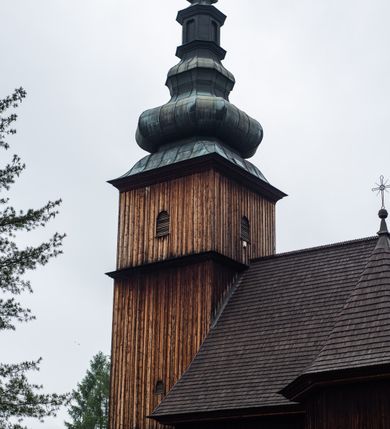 Zdjęcie nr 28: Kościół drewniany o konstrukcji zrębowej, elewacje jednolite, szalowane z listwowaniem, wzmocnione lisicami, wieża o konstrukcji słupowej. Więźba dachowa drewniana, krokwiowa. Budynek na kamiennym podmurowaniu przesłoniętym daszkiem gontowym. Kościół orientowany, jednonawowy z dwiema kaplicami o charakterze transeptu zamkniętymi wielobocznie, na planie krzyża łacińskiego, o węższym prezbiterium zamkniętym trójbocznie. Do wschodniej ściany prezbiterium przylega ogrójec w formie kapliczki, od północy zakrystia na planie prostokąta. Na skrzyżowaniu nawy z południowym ramieniem transeptu kruchta na planie prostokąta. Od zachodu do nawy przylega wieża na planie kwadratu, otoczona sobotami. Prezbiterium i nawa nakryte pozornym sklepieniem tej samej szerokości, obwiedzionym dużym, profilowanym gzymsem podtrzymywanym w nawie przez dwie pary filarów o kanelowanych trzonach, na cokołach. Sklepieniu nawy głównej towarzyszą płaskie i niższe stropy z fasetami w przestrzeniach bocznych. Ściany boczne nawy zwieńczone profilowanym gzymsem. Pomiędzy nawą a prezbiterium otwór tęczowy w formie odcinkowej arkady podtrzymywanej przez filary o kanelowanych trzonach z belką o falistym zarysie. Kaplice boczne otwarte do nawy odcinkowymi arkadami podtrzymywanymi przez filary o kanelowanych trzonach, na cokołach. Parapet chóru muzycznego w zachodniej części nawy, o wybrzuszonej środkowej części, podparty na dwóch filarach o kanelowanych trzonach, na cokołach. Posadzka z płyt ceramicznych. Elewacja frontowa w postaci wieży otoczonej sobotami. Na osi frontowej ściany sobót płytka kruchta zwieńczona gzymsem z przyczółkiem wypełnionym motywem „słoneczka”, przykryta dwuspadowym dachem. Drzwi dwuskrzydłowe, drewniane z okuciami. Korpus wieży podzielony poziomo daszkiem okapowym w dwóch trzecich wysokości, na froncie okno. W dolnej części małe otwory dzwonne z trzech stron, w górnej części duże otwory dzwonne z czterech stron. Wieża zwieńczona hełmem baniastym z latarnią. Ściany sobót szalowane z poziomym listwowaniem, przekryte mansardowym dachem gontowym, na północnej i południowej ścianie dwie pary okien w formie leżącego prostokąta zamkniętego łukiem odcinkowym. W elewacji południowej cztery otwory okienne w formie stojącego prostokąta zamkniętego łukiem odcinkowym, w partii prezbiterium z witrażami. Na elewacji wschodniej ogrójec w formie nadwieszonej kapliczki nakrytej dwuspadowym daszkiem z grupą Ukrzyżowania. W elewacji północnej dwa otwory okienne w formie stojącego prostokąta zamkniętego łukiem odcinkowym. W zakrystii małe kwadratowe okienko. W obu ramionach transeptu analogiczne jak w elewacjach otwory okienne na wschodniej i zachodniej ścianie. Kościół nakryty dachami gontowymi, wyższym nad nawą, niższym nad prezbiterium, dwuspadowym nad nawą oraz wielospadowym nad prezbiterium i ramionami transeptu; nad zakrystią i kruchtą boczną dachy dwuspadowe. W połowie długości dachu, nad nawą, od strony prezbiterium wznosi się ponad kalenicą wieżyczka na sygnaturkę, obita blachą, na ośmiobocznym trzonie, z latarnią zwieńczoną niewielkim baniastym hełmem z latarnią.
