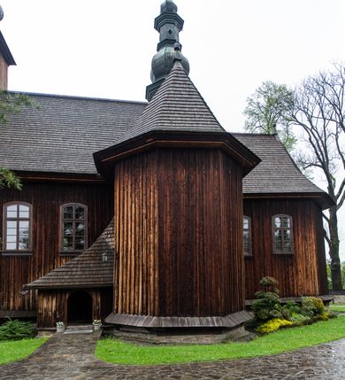 Zdjęcie nr 36: Kościół drewniany o konstrukcji zrębowej, elewacje jednolite, szalowane z listwowaniem, wzmocnione lisicami, wieża o konstrukcji słupowej. Więźba dachowa drewniana, krokwiowa. Budynek na kamiennym podmurowaniu przesłoniętym daszkiem gontowym. Kościół orientowany, jednonawowy z dwiema kaplicami o charakterze transeptu zamkniętymi wielobocznie, na planie krzyża łacińskiego, o węższym prezbiterium zamkniętym trójbocznie. Do wschodniej ściany prezbiterium przylega ogrójec w formie kapliczki, od północy zakrystia na planie prostokąta. Na skrzyżowaniu nawy z południowym ramieniem transeptu kruchta na planie prostokąta. Od zachodu do nawy przylega wieża na planie kwadratu, otoczona sobotami. Prezbiterium i nawa nakryte pozornym sklepieniem tej samej szerokości, obwiedzionym dużym, profilowanym gzymsem podtrzymywanym w nawie przez dwie pary filarów o kanelowanych trzonach, na cokołach. Sklepieniu nawy głównej towarzyszą płaskie i niższe stropy z fasetami w przestrzeniach bocznych. Ściany boczne nawy zwieńczone profilowanym gzymsem. Pomiędzy nawą a prezbiterium otwór tęczowy w formie odcinkowej arkady podtrzymywanej przez filary o kanelowanych trzonach z belką o falistym zarysie. Kaplice boczne otwarte do nawy odcinkowymi arkadami podtrzymywanymi przez filary o kanelowanych trzonach, na cokołach. Parapet chóru muzycznego w zachodniej części nawy, o wybrzuszonej środkowej części, podparty na dwóch filarach o kanelowanych trzonach, na cokołach. Posadzka z płyt ceramicznych. Elewacja frontowa w postaci wieży otoczonej sobotami. Na osi frontowej ściany sobót płytka kruchta zwieńczona gzymsem z przyczółkiem wypełnionym motywem „słoneczka”, przykryta dwuspadowym dachem. Drzwi dwuskrzydłowe, drewniane z okuciami. Korpus wieży podzielony poziomo daszkiem okapowym w dwóch trzecich wysokości, na froncie okno. W dolnej części małe otwory dzwonne z trzech stron, w górnej części duże otwory dzwonne z czterech stron. Wieża zwieńczona hełmem baniastym z latarnią. Ściany sobót szalowane z poziomym listwowaniem, przekryte mansardowym dachem gontowym, na północnej i południowej ścianie dwie pary okien w formie leżącego prostokąta zamkniętego łukiem odcinkowym. W elewacji południowej cztery otwory okienne w formie stojącego prostokąta zamkniętego łukiem odcinkowym, w partii prezbiterium z witrażami. Na elewacji wschodniej ogrójec w formie nadwieszonej kapliczki nakrytej dwuspadowym daszkiem z grupą Ukrzyżowania. W elewacji północnej dwa otwory okienne w formie stojącego prostokąta zamkniętego łukiem odcinkowym. W zakrystii małe kwadratowe okienko. W obu ramionach transeptu analogiczne jak w elewacjach otwory okienne na wschodniej i zachodniej ścianie. Kościół nakryty dachami gontowymi, wyższym nad nawą, niższym nad prezbiterium, dwuspadowym nad nawą oraz wielospadowym nad prezbiterium i ramionami transeptu; nad zakrystią i kruchtą boczną dachy dwuspadowe. W połowie długości dachu, nad nawą, od strony prezbiterium wznosi się ponad kalenicą wieżyczka na sygnaturkę, obita blachą, na ośmiobocznym trzonie, z latarnią zwieńczoną niewielkim baniastym hełmem z latarnią.
