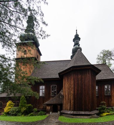 Zdjęcie nr 34: Kościół drewniany o konstrukcji zrębowej, elewacje jednolite, szalowane z listwowaniem, wzmocnione lisicami, wieża o konstrukcji słupowej. Więźba dachowa drewniana, krokwiowa. Budynek na kamiennym podmurowaniu przesłoniętym daszkiem gontowym. Kościół orientowany, jednonawowy z dwiema kaplicami o charakterze transeptu zamkniętymi wielobocznie, na planie krzyża łacińskiego, o węższym prezbiterium zamkniętym trójbocznie. Do wschodniej ściany prezbiterium przylega ogrójec w formie kapliczki, od północy zakrystia na planie prostokąta. Na skrzyżowaniu nawy z południowym ramieniem transeptu kruchta na planie prostokąta. Od zachodu do nawy przylega wieża na planie kwadratu, otoczona sobotami. Prezbiterium i nawa nakryte pozornym sklepieniem tej samej szerokości, obwiedzionym dużym, profilowanym gzymsem podtrzymywanym w nawie przez dwie pary filarów o kanelowanych trzonach, na cokołach. Sklepieniu nawy głównej towarzyszą płaskie i niższe stropy z fasetami w przestrzeniach bocznych. Ściany boczne nawy zwieńczone profilowanym gzymsem. Pomiędzy nawą a prezbiterium otwór tęczowy w formie odcinkowej arkady podtrzymywanej przez filary o kanelowanych trzonach z belką o falistym zarysie. Kaplice boczne otwarte do nawy odcinkowymi arkadami podtrzymywanymi przez filary o kanelowanych trzonach, na cokołach. Parapet chóru muzycznego w zachodniej części nawy, o wybrzuszonej środkowej części, podparty na dwóch filarach o kanelowanych trzonach, na cokołach. Posadzka z płyt ceramicznych. Elewacja frontowa w postaci wieży otoczonej sobotami. Na osi frontowej ściany sobót płytka kruchta zwieńczona gzymsem z przyczółkiem wypełnionym motywem „słoneczka”, przykryta dwuspadowym dachem. Drzwi dwuskrzydłowe, drewniane z okuciami. Korpus wieży podzielony poziomo daszkiem okapowym w dwóch trzecich wysokości, na froncie okno. W dolnej części małe otwory dzwonne z trzech stron, w górnej części duże otwory dzwonne z czterech stron. Wieża zwieńczona hełmem baniastym z latarnią. Ściany sobót szalowane z poziomym listwowaniem, przekryte mansardowym dachem gontowym, na północnej i południowej ścianie dwie pary okien w formie leżącego prostokąta zamkniętego łukiem odcinkowym. W elewacji południowej cztery otwory okienne w formie stojącego prostokąta zamkniętego łukiem odcinkowym, w partii prezbiterium z witrażami. Na elewacji wschodniej ogrójec w formie nadwieszonej kapliczki nakrytej dwuspadowym daszkiem z grupą Ukrzyżowania. W elewacji północnej dwa otwory okienne w formie stojącego prostokąta zamkniętego łukiem odcinkowym. W zakrystii małe kwadratowe okienko. W obu ramionach transeptu analogiczne jak w elewacjach otwory okienne na wschodniej i zachodniej ścianie. Kościół nakryty dachami gontowymi, wyższym nad nawą, niższym nad prezbiterium, dwuspadowym nad nawą oraz wielospadowym nad prezbiterium i ramionami transeptu; nad zakrystią i kruchtą boczną dachy dwuspadowe. W połowie długości dachu, nad nawą, od strony prezbiterium wznosi się ponad kalenicą wieżyczka na sygnaturkę, obita blachą, na ośmiobocznym trzonie, z latarnią zwieńczoną niewielkim baniastym hełmem z latarnią.
