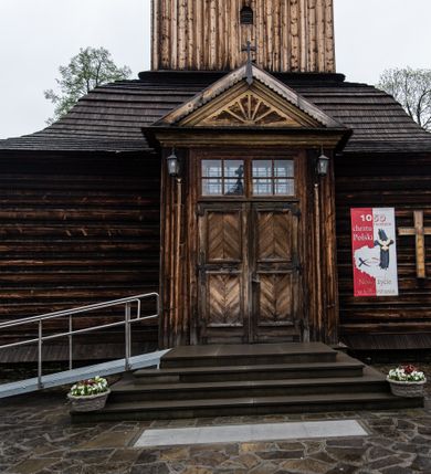 Zdjęcie nr 41: Kościół drewniany o konstrukcji zrębowej, elewacje jednolite, szalowane z listwowaniem, wzmocnione lisicami, wieża o konstrukcji słupowej. Więźba dachowa drewniana, krokwiowa. Budynek na kamiennym podmurowaniu przesłoniętym daszkiem gontowym. Kościół orientowany, jednonawowy z dwiema kaplicami o charakterze transeptu zamkniętymi wielobocznie, na planie krzyża łacińskiego, o węższym prezbiterium zamkniętym trójbocznie. Do wschodniej ściany prezbiterium przylega ogrójec w formie kapliczki, od północy zakrystia na planie prostokąta. Na skrzyżowaniu nawy z południowym ramieniem transeptu kruchta na planie prostokąta. Od zachodu do nawy przylega wieża na planie kwadratu, otoczona sobotami. Prezbiterium i nawa nakryte pozornym sklepieniem tej samej szerokości, obwiedzionym dużym, profilowanym gzymsem podtrzymywanym w nawie przez dwie pary filarów o kanelowanych trzonach, na cokołach. Sklepieniu nawy głównej towarzyszą płaskie i niższe stropy z fasetami w przestrzeniach bocznych. Ściany boczne nawy zwieńczone profilowanym gzymsem. Pomiędzy nawą a prezbiterium otwór tęczowy w formie odcinkowej arkady podtrzymywanej przez filary o kanelowanych trzonach z belką o falistym zarysie. Kaplice boczne otwarte do nawy odcinkowymi arkadami podtrzymywanymi przez filary o kanelowanych trzonach, na cokołach. Parapet chóru muzycznego w zachodniej części nawy, o wybrzuszonej środkowej części, podparty na dwóch filarach o kanelowanych trzonach, na cokołach. Posadzka z płyt ceramicznych. Elewacja frontowa w postaci wieży otoczonej sobotami. Na osi frontowej ściany sobót płytka kruchta zwieńczona gzymsem z przyczółkiem wypełnionym motywem „słoneczka”, przykryta dwuspadowym dachem. Drzwi dwuskrzydłowe, drewniane z okuciami. Korpus wieży podzielony poziomo daszkiem okapowym w dwóch trzecich wysokości, na froncie okno. W dolnej części małe otwory dzwonne z trzech stron, w górnej części duże otwory dzwonne z czterech stron. Wieża zwieńczona hełmem baniastym z latarnią. Ściany sobót szalowane z poziomym listwowaniem, przekryte mansardowym dachem gontowym, na północnej i południowej ścianie dwie pary okien w formie leżącego prostokąta zamkniętego łukiem odcinkowym. W elewacji południowej cztery otwory okienne w formie stojącego prostokąta zamkniętego łukiem odcinkowym, w partii prezbiterium z witrażami. Na elewacji wschodniej ogrójec w formie nadwieszonej kapliczki nakrytej dwuspadowym daszkiem z grupą Ukrzyżowania. W elewacji północnej dwa otwory okienne w formie stojącego prostokąta zamkniętego łukiem odcinkowym. W zakrystii małe kwadratowe okienko. W obu ramionach transeptu analogiczne jak w elewacjach otwory okienne na wschodniej i zachodniej ścianie. Kościół nakryty dachami gontowymi, wyższym nad nawą, niższym nad prezbiterium, dwuspadowym nad nawą oraz wielospadowym nad prezbiterium i ramionami transeptu; nad zakrystią i kruchtą boczną dachy dwuspadowe. W połowie długości dachu, nad nawą, od strony prezbiterium wznosi się ponad kalenicą wieżyczka na sygnaturkę, obita blachą, na ośmiobocznym trzonie, z latarnią zwieńczoną niewielkim baniastym hełmem z latarnią.
