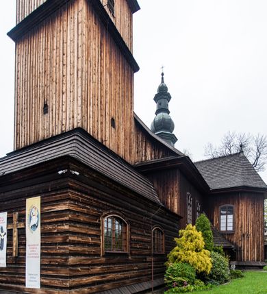 Zdjęcie nr 39: Kościół drewniany o konstrukcji zrębowej, elewacje jednolite, szalowane z listwowaniem, wzmocnione lisicami, wieża o konstrukcji słupowej. Więźba dachowa drewniana, krokwiowa. Budynek na kamiennym podmurowaniu przesłoniętym daszkiem gontowym. Kościół orientowany, jednonawowy z dwiema kaplicami o charakterze transeptu zamkniętymi wielobocznie, na planie krzyża łacińskiego, o węższym prezbiterium zamkniętym trójbocznie. Do wschodniej ściany prezbiterium przylega ogrójec w formie kapliczki, od północy zakrystia na planie prostokąta. Na skrzyżowaniu nawy z południowym ramieniem transeptu kruchta na planie prostokąta. Od zachodu do nawy przylega wieża na planie kwadratu, otoczona sobotami. Prezbiterium i nawa nakryte pozornym sklepieniem tej samej szerokości, obwiedzionym dużym, profilowanym gzymsem podtrzymywanym w nawie przez dwie pary filarów o kanelowanych trzonach, na cokołach. Sklepieniu nawy głównej towarzyszą płaskie i niższe stropy z fasetami w przestrzeniach bocznych. Ściany boczne nawy zwieńczone profilowanym gzymsem. Pomiędzy nawą a prezbiterium otwór tęczowy w formie odcinkowej arkady podtrzymywanej przez filary o kanelowanych trzonach z belką o falistym zarysie. Kaplice boczne otwarte do nawy odcinkowymi arkadami podtrzymywanymi przez filary o kanelowanych trzonach, na cokołach. Parapet chóru muzycznego w zachodniej części nawy, o wybrzuszonej środkowej części, podparty na dwóch filarach o kanelowanych trzonach, na cokołach. Posadzka z płyt ceramicznych. Elewacja frontowa w postaci wieży otoczonej sobotami. Na osi frontowej ściany sobót płytka kruchta zwieńczona gzymsem z przyczółkiem wypełnionym motywem „słoneczka”, przykryta dwuspadowym dachem. Drzwi dwuskrzydłowe, drewniane z okuciami. Korpus wieży podzielony poziomo daszkiem okapowym w dwóch trzecich wysokości, na froncie okno. W dolnej części małe otwory dzwonne z trzech stron, w górnej części duże otwory dzwonne z czterech stron. Wieża zwieńczona hełmem baniastym z latarnią. Ściany sobót szalowane z poziomym listwowaniem, przekryte mansardowym dachem gontowym, na północnej i południowej ścianie dwie pary okien w formie leżącego prostokąta zamkniętego łukiem odcinkowym. W elewacji południowej cztery otwory okienne w formie stojącego prostokąta zamkniętego łukiem odcinkowym, w partii prezbiterium z witrażami. Na elewacji wschodniej ogrójec w formie nadwieszonej kapliczki nakrytej dwuspadowym daszkiem z grupą Ukrzyżowania. W elewacji północnej dwa otwory okienne w formie stojącego prostokąta zamkniętego łukiem odcinkowym. W zakrystii małe kwadratowe okienko. W obu ramionach transeptu analogiczne jak w elewacjach otwory okienne na wschodniej i zachodniej ścianie. Kościół nakryty dachami gontowymi, wyższym nad nawą, niższym nad prezbiterium, dwuspadowym nad nawą oraz wielospadowym nad prezbiterium i ramionami transeptu; nad zakrystią i kruchtą boczną dachy dwuspadowe. W połowie długości dachu, nad nawą, od strony prezbiterium wznosi się ponad kalenicą wieżyczka na sygnaturkę, obita blachą, na ośmiobocznym trzonie, z latarnią zwieńczoną niewielkim baniastym hełmem z latarnią.
