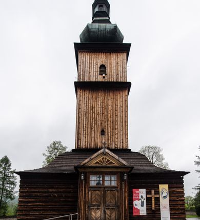 Zdjęcie nr 40: Kościół drewniany o konstrukcji zrębowej, elewacje jednolite, szalowane z listwowaniem, wzmocnione lisicami, wieża o konstrukcji słupowej. Więźba dachowa drewniana, krokwiowa. Budynek na kamiennym podmurowaniu przesłoniętym daszkiem gontowym. Kościół orientowany, jednonawowy z dwiema kaplicami o charakterze transeptu zamkniętymi wielobocznie, na planie krzyża łacińskiego, o węższym prezbiterium zamkniętym trójbocznie. Do wschodniej ściany prezbiterium przylega ogrójec w formie kapliczki, od północy zakrystia na planie prostokąta. Na skrzyżowaniu nawy z południowym ramieniem transeptu kruchta na planie prostokąta. Od zachodu do nawy przylega wieża na planie kwadratu, otoczona sobotami. Prezbiterium i nawa nakryte pozornym sklepieniem tej samej szerokości, obwiedzionym dużym, profilowanym gzymsem podtrzymywanym w nawie przez dwie pary filarów o kanelowanych trzonach, na cokołach. Sklepieniu nawy głównej towarzyszą płaskie i niższe stropy z fasetami w przestrzeniach bocznych. Ściany boczne nawy zwieńczone profilowanym gzymsem. Pomiędzy nawą a prezbiterium otwór tęczowy w formie odcinkowej arkady podtrzymywanej przez filary o kanelowanych trzonach z belką o falistym zarysie. Kaplice boczne otwarte do nawy odcinkowymi arkadami podtrzymywanymi przez filary o kanelowanych trzonach, na cokołach. Parapet chóru muzycznego w zachodniej części nawy, o wybrzuszonej środkowej części, podparty na dwóch filarach o kanelowanych trzonach, na cokołach. Posadzka z płyt ceramicznych. Elewacja frontowa w postaci wieży otoczonej sobotami. Na osi frontowej ściany sobót płytka kruchta zwieńczona gzymsem z przyczółkiem wypełnionym motywem „słoneczka”, przykryta dwuspadowym dachem. Drzwi dwuskrzydłowe, drewniane z okuciami. Korpus wieży podzielony poziomo daszkiem okapowym w dwóch trzecich wysokości, na froncie okno. W dolnej części małe otwory dzwonne z trzech stron, w górnej części duże otwory dzwonne z czterech stron. Wieża zwieńczona hełmem baniastym z latarnią. Ściany sobót szalowane z poziomym listwowaniem, przekryte mansardowym dachem gontowym, na północnej i południowej ścianie dwie pary okien w formie leżącego prostokąta zamkniętego łukiem odcinkowym. W elewacji południowej cztery otwory okienne w formie stojącego prostokąta zamkniętego łukiem odcinkowym, w partii prezbiterium z witrażami. Na elewacji wschodniej ogrójec w formie nadwieszonej kapliczki nakrytej dwuspadowym daszkiem z grupą Ukrzyżowania. W elewacji północnej dwa otwory okienne w formie stojącego prostokąta zamkniętego łukiem odcinkowym. W zakrystii małe kwadratowe okienko. W obu ramionach transeptu analogiczne jak w elewacjach otwory okienne na wschodniej i zachodniej ścianie. Kościół nakryty dachami gontowymi, wyższym nad nawą, niższym nad prezbiterium, dwuspadowym nad nawą oraz wielospadowym nad prezbiterium i ramionami transeptu; nad zakrystią i kruchtą boczną dachy dwuspadowe. W połowie długości dachu, nad nawą, od strony prezbiterium wznosi się ponad kalenicą wieżyczka na sygnaturkę, obita blachą, na ośmiobocznym trzonie, z latarnią zwieńczoną niewielkim baniastym hełmem z latarnią.
