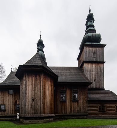 Zdjęcie nr 49: Kościół drewniany o konstrukcji zrębowej, elewacje jednolite, szalowane z listwowaniem, wzmocnione lisicami, wieża o konstrukcji słupowej. Więźba dachowa drewniana, krokwiowa. Budynek na kamiennym podmurowaniu przesłoniętym daszkiem gontowym. Kościół orientowany, jednonawowy z dwiema kaplicami o charakterze transeptu zamkniętymi wielobocznie, na planie krzyża łacińskiego, o węższym prezbiterium zamkniętym trójbocznie. Do wschodniej ściany prezbiterium przylega ogrójec w formie kapliczki, od północy zakrystia na planie prostokąta. Na skrzyżowaniu nawy z południowym ramieniem transeptu kruchta na planie prostokąta. Od zachodu do nawy przylega wieża na planie kwadratu, otoczona sobotami. Prezbiterium i nawa nakryte pozornym sklepieniem tej samej szerokości, obwiedzionym dużym, profilowanym gzymsem podtrzymywanym w nawie przez dwie pary filarów o kanelowanych trzonach, na cokołach. Sklepieniu nawy głównej towarzyszą płaskie i niższe stropy z fasetami w przestrzeniach bocznych. Ściany boczne nawy zwieńczone profilowanym gzymsem. Pomiędzy nawą a prezbiterium otwór tęczowy w formie odcinkowej arkady podtrzymywanej przez filary o kanelowanych trzonach z belką o falistym zarysie. Kaplice boczne otwarte do nawy odcinkowymi arkadami podtrzymywanymi przez filary o kanelowanych trzonach, na cokołach. Parapet chóru muzycznego w zachodniej części nawy, o wybrzuszonej środkowej części, podparty na dwóch filarach o kanelowanych trzonach, na cokołach. Posadzka z płyt ceramicznych. Elewacja frontowa w postaci wieży otoczonej sobotami. Na osi frontowej ściany sobót płytka kruchta zwieńczona gzymsem z przyczółkiem wypełnionym motywem „słoneczka”, przykryta dwuspadowym dachem. Drzwi dwuskrzydłowe, drewniane z okuciami. Korpus wieży podzielony poziomo daszkiem okapowym w dwóch trzecich wysokości, na froncie okno. W dolnej części małe otwory dzwonne z trzech stron, w górnej części duże otwory dzwonne z czterech stron. Wieża zwieńczona hełmem baniastym z latarnią. Ściany sobót szalowane z poziomym listwowaniem, przekryte mansardowym dachem gontowym, na północnej i południowej ścianie dwie pary okien w formie leżącego prostokąta zamkniętego łukiem odcinkowym. W elewacji południowej cztery otwory okienne w formie stojącego prostokąta zamkniętego łukiem odcinkowym, w partii prezbiterium z witrażami. Na elewacji wschodniej ogrójec w formie nadwieszonej kapliczki nakrytej dwuspadowym daszkiem z grupą Ukrzyżowania. W elewacji północnej dwa otwory okienne w formie stojącego prostokąta zamkniętego łukiem odcinkowym. W zakrystii małe kwadratowe okienko. W obu ramionach transeptu analogiczne jak w elewacjach otwory okienne na wschodniej i zachodniej ścianie. Kościół nakryty dachami gontowymi, wyższym nad nawą, niższym nad prezbiterium, dwuspadowym nad nawą oraz wielospadowym nad prezbiterium i ramionami transeptu; nad zakrystią i kruchtą boczną dachy dwuspadowe. W połowie długości dachu, nad nawą, od strony prezbiterium wznosi się ponad kalenicą wieżyczka na sygnaturkę, obita blachą, na ośmiobocznym trzonie, z latarnią zwieńczoną niewielkim baniastym hełmem z latarnią.
