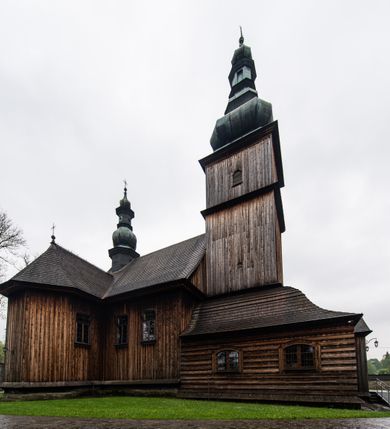 Zdjęcie nr 46: Kościół drewniany o konstrukcji zrębowej, elewacje jednolite, szalowane z listwowaniem, wzmocnione lisicami, wieża o konstrukcji słupowej. Więźba dachowa drewniana, krokwiowa. Budynek na kamiennym podmurowaniu przesłoniętym daszkiem gontowym. Kościół orientowany, jednonawowy z dwiema kaplicami o charakterze transeptu zamkniętymi wielobocznie, na planie krzyża łacińskiego, o węższym prezbiterium zamkniętym trójbocznie. Do wschodniej ściany prezbiterium przylega ogrójec w formie kapliczki, od północy zakrystia na planie prostokąta. Na skrzyżowaniu nawy z południowym ramieniem transeptu kruchta na planie prostokąta. Od zachodu do nawy przylega wieża na planie kwadratu, otoczona sobotami. Prezbiterium i nawa nakryte pozornym sklepieniem tej samej szerokości, obwiedzionym dużym, profilowanym gzymsem podtrzymywanym w nawie przez dwie pary filarów o kanelowanych trzonach, na cokołach. Sklepieniu nawy głównej towarzyszą płaskie i niższe stropy z fasetami w przestrzeniach bocznych. Ściany boczne nawy zwieńczone profilowanym gzymsem. Pomiędzy nawą a prezbiterium otwór tęczowy w formie odcinkowej arkady podtrzymywanej przez filary o kanelowanych trzonach z belką o falistym zarysie. Kaplice boczne otwarte do nawy odcinkowymi arkadami podtrzymywanymi przez filary o kanelowanych trzonach, na cokołach. Parapet chóru muzycznego w zachodniej części nawy, o wybrzuszonej środkowej części, podparty na dwóch filarach o kanelowanych trzonach, na cokołach. Posadzka z płyt ceramicznych. Elewacja frontowa w postaci wieży otoczonej sobotami. Na osi frontowej ściany sobót płytka kruchta zwieńczona gzymsem z przyczółkiem wypełnionym motywem „słoneczka”, przykryta dwuspadowym dachem. Drzwi dwuskrzydłowe, drewniane z okuciami. Korpus wieży podzielony poziomo daszkiem okapowym w dwóch trzecich wysokości, na froncie okno. W dolnej części małe otwory dzwonne z trzech stron, w górnej części duże otwory dzwonne z czterech stron. Wieża zwieńczona hełmem baniastym z latarnią. Ściany sobót szalowane z poziomym listwowaniem, przekryte mansardowym dachem gontowym, na północnej i południowej ścianie dwie pary okien w formie leżącego prostokąta zamkniętego łukiem odcinkowym. W elewacji południowej cztery otwory okienne w formie stojącego prostokąta zamkniętego łukiem odcinkowym, w partii prezbiterium z witrażami. Na elewacji wschodniej ogrójec w formie nadwieszonej kapliczki nakrytej dwuspadowym daszkiem z grupą Ukrzyżowania. W elewacji północnej dwa otwory okienne w formie stojącego prostokąta zamkniętego łukiem odcinkowym. W zakrystii małe kwadratowe okienko. W obu ramionach transeptu analogiczne jak w elewacjach otwory okienne na wschodniej i zachodniej ścianie. Kościół nakryty dachami gontowymi, wyższym nad nawą, niższym nad prezbiterium, dwuspadowym nad nawą oraz wielospadowym nad prezbiterium i ramionami transeptu; nad zakrystią i kruchtą boczną dachy dwuspadowe. W połowie długości dachu, nad nawą, od strony prezbiterium wznosi się ponad kalenicą wieżyczka na sygnaturkę, obita blachą, na ośmiobocznym trzonie, z latarnią zwieńczoną niewielkim baniastym hełmem z latarnią.
