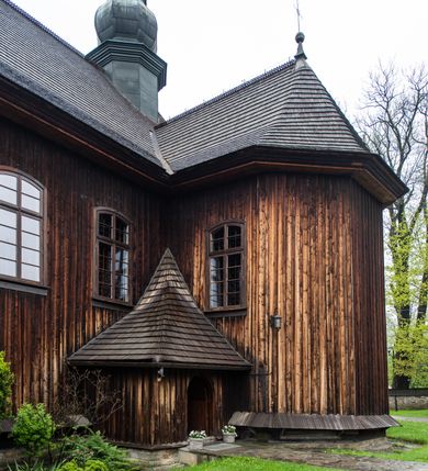 Zdjęcie nr 35: Kościół drewniany o konstrukcji zrębowej, elewacje jednolite, szalowane z listwowaniem, wzmocnione lisicami, wieża o konstrukcji słupowej. Więźba dachowa drewniana, krokwiowa. Budynek na kamiennym podmurowaniu przesłoniętym daszkiem gontowym. Kościół orientowany, jednonawowy z dwiema kaplicami o charakterze transeptu zamkniętymi wielobocznie, na planie krzyża łacińskiego, o węższym prezbiterium zamkniętym trójbocznie. Do wschodniej ściany prezbiterium przylega ogrójec w formie kapliczki, od północy zakrystia na planie prostokąta. Na skrzyżowaniu nawy z południowym ramieniem transeptu kruchta na planie prostokąta. Od zachodu do nawy przylega wieża na planie kwadratu, otoczona sobotami. Prezbiterium i nawa nakryte pozornym sklepieniem tej samej szerokości, obwiedzionym dużym, profilowanym gzymsem podtrzymywanym w nawie przez dwie pary filarów o kanelowanych trzonach, na cokołach. Sklepieniu nawy głównej towarzyszą płaskie i niższe stropy z fasetami w przestrzeniach bocznych. Ściany boczne nawy zwieńczone profilowanym gzymsem. Pomiędzy nawą a prezbiterium otwór tęczowy w formie odcinkowej arkady podtrzymywanej przez filary o kanelowanych trzonach z belką o falistym zarysie. Kaplice boczne otwarte do nawy odcinkowymi arkadami podtrzymywanymi przez filary o kanelowanych trzonach, na cokołach. Parapet chóru muzycznego w zachodniej części nawy, o wybrzuszonej środkowej części, podparty na dwóch filarach o kanelowanych trzonach, na cokołach. Posadzka z płyt ceramicznych. Elewacja frontowa w postaci wieży otoczonej sobotami. Na osi frontowej ściany sobót płytka kruchta zwieńczona gzymsem z przyczółkiem wypełnionym motywem „słoneczka”, przykryta dwuspadowym dachem. Drzwi dwuskrzydłowe, drewniane z okuciami. Korpus wieży podzielony poziomo daszkiem okapowym w dwóch trzecich wysokości, na froncie okno. W dolnej części małe otwory dzwonne z trzech stron, w górnej części duże otwory dzwonne z czterech stron. Wieża zwieńczona hełmem baniastym z latarnią. Ściany sobót szalowane z poziomym listwowaniem, przekryte mansardowym dachem gontowym, na północnej i południowej ścianie dwie pary okien w formie leżącego prostokąta zamkniętego łukiem odcinkowym. W elewacji południowej cztery otwory okienne w formie stojącego prostokąta zamkniętego łukiem odcinkowym, w partii prezbiterium z witrażami. Na elewacji wschodniej ogrójec w formie nadwieszonej kapliczki nakrytej dwuspadowym daszkiem z grupą Ukrzyżowania. W elewacji północnej dwa otwory okienne w formie stojącego prostokąta zamkniętego łukiem odcinkowym. W zakrystii małe kwadratowe okienko. W obu ramionach transeptu analogiczne jak w elewacjach otwory okienne na wschodniej i zachodniej ścianie. Kościół nakryty dachami gontowymi, wyższym nad nawą, niższym nad prezbiterium, dwuspadowym nad nawą oraz wielospadowym nad prezbiterium i ramionami transeptu; nad zakrystią i kruchtą boczną dachy dwuspadowe. W połowie długości dachu, nad nawą, od strony prezbiterium wznosi się ponad kalenicą wieżyczka na sygnaturkę, obita blachą, na ośmiobocznym trzonie, z latarnią zwieńczoną niewielkim baniastym hełmem z latarnią.
