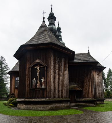 Zdjęcie nr 55: Kościół drewniany o konstrukcji zrębowej, elewacje jednolite, szalowane z listwowaniem, wzmocnione lisicami, wieża o konstrukcji słupowej. Więźba dachowa drewniana, krokwiowa. Budynek na kamiennym podmurowaniu przesłoniętym daszkiem gontowym. Kościół orientowany, jednonawowy z dwiema kaplicami o charakterze transeptu zamkniętymi wielobocznie, na planie krzyża łacińskiego, o węższym prezbiterium zamkniętym trójbocznie. Do wschodniej ściany prezbiterium przylega ogrójec w formie kapliczki, od północy zakrystia na planie prostokąta. Na skrzyżowaniu nawy z południowym ramieniem transeptu kruchta na planie prostokąta. Od zachodu do nawy przylega wieża na planie kwadratu, otoczona sobotami. Prezbiterium i nawa nakryte pozornym sklepieniem tej samej szerokości, obwiedzionym dużym, profilowanym gzymsem podtrzymywanym w nawie przez dwie pary filarów o kanelowanych trzonach, na cokołach. Sklepieniu nawy głównej towarzyszą płaskie i niższe stropy z fasetami w przestrzeniach bocznych. Ściany boczne nawy zwieńczone profilowanym gzymsem. Pomiędzy nawą a prezbiterium otwór tęczowy w formie odcinkowej arkady podtrzymywanej przez filary o kanelowanych trzonach z belką o falistym zarysie. Kaplice boczne otwarte do nawy odcinkowymi arkadami podtrzymywanymi przez filary o kanelowanych trzonach, na cokołach. Parapet chóru muzycznego w zachodniej części nawy, o wybrzuszonej środkowej części, podparty na dwóch filarach o kanelowanych trzonach, na cokołach. Posadzka z płyt ceramicznych. Elewacja frontowa w postaci wieży otoczonej sobotami. Na osi frontowej ściany sobót płytka kruchta zwieńczona gzymsem z przyczółkiem wypełnionym motywem „słoneczka”, przykryta dwuspadowym dachem. Drzwi dwuskrzydłowe, drewniane z okuciami. Korpus wieży podzielony poziomo daszkiem okapowym w dwóch trzecich wysokości, na froncie okno. W dolnej części małe otwory dzwonne z trzech stron, w górnej części duże otwory dzwonne z czterech stron. Wieża zwieńczona hełmem baniastym z latarnią. Ściany sobót szalowane z poziomym listwowaniem, przekryte mansardowym dachem gontowym, na północnej i południowej ścianie dwie pary okien w formie leżącego prostokąta zamkniętego łukiem odcinkowym. W elewacji południowej cztery otwory okienne w formie stojącego prostokąta zamkniętego łukiem odcinkowym, w partii prezbiterium z witrażami. Na elewacji wschodniej ogrójec w formie nadwieszonej kapliczki nakrytej dwuspadowym daszkiem z grupą Ukrzyżowania. W elewacji północnej dwa otwory okienne w formie stojącego prostokąta zamkniętego łukiem odcinkowym. W zakrystii małe kwadratowe okienko. W obu ramionach transeptu analogiczne jak w elewacjach otwory okienne na wschodniej i zachodniej ścianie. Kościół nakryty dachami gontowymi, wyższym nad nawą, niższym nad prezbiterium, dwuspadowym nad nawą oraz wielospadowym nad prezbiterium i ramionami transeptu; nad zakrystią i kruchtą boczną dachy dwuspadowe. W połowie długości dachu, nad nawą, od strony prezbiterium wznosi się ponad kalenicą wieżyczka na sygnaturkę, obita blachą, na ośmiobocznym trzonie, z latarnią zwieńczoną niewielkim baniastym hełmem z latarnią.
