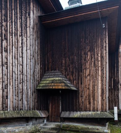 Zdjęcie nr 52: Kościół drewniany o konstrukcji zrębowej, elewacje jednolite, szalowane z listwowaniem, wzmocnione lisicami, wieża o konstrukcji słupowej. Więźba dachowa drewniana, krokwiowa. Budynek na kamiennym podmurowaniu przesłoniętym daszkiem gontowym. Kościół orientowany, jednonawowy z dwiema kaplicami o charakterze transeptu zamkniętymi wielobocznie, na planie krzyża łacińskiego, o węższym prezbiterium zamkniętym trójbocznie. Do wschodniej ściany prezbiterium przylega ogrójec w formie kapliczki, od północy zakrystia na planie prostokąta. Na skrzyżowaniu nawy z południowym ramieniem transeptu kruchta na planie prostokąta. Od zachodu do nawy przylega wieża na planie kwadratu, otoczona sobotami. Prezbiterium i nawa nakryte pozornym sklepieniem tej samej szerokości, obwiedzionym dużym, profilowanym gzymsem podtrzymywanym w nawie przez dwie pary filarów o kanelowanych trzonach, na cokołach. Sklepieniu nawy głównej towarzyszą płaskie i niższe stropy z fasetami w przestrzeniach bocznych. Ściany boczne nawy zwieńczone profilowanym gzymsem. Pomiędzy nawą a prezbiterium otwór tęczowy w formie odcinkowej arkady podtrzymywanej przez filary o kanelowanych trzonach z belką o falistym zarysie. Kaplice boczne otwarte do nawy odcinkowymi arkadami podtrzymywanymi przez filary o kanelowanych trzonach, na cokołach. Parapet chóru muzycznego w zachodniej części nawy, o wybrzuszonej środkowej części, podparty na dwóch filarach o kanelowanych trzonach, na cokołach. Posadzka z płyt ceramicznych. Elewacja frontowa w postaci wieży otoczonej sobotami. Na osi frontowej ściany sobót płytka kruchta zwieńczona gzymsem z przyczółkiem wypełnionym motywem „słoneczka”, przykryta dwuspadowym dachem. Drzwi dwuskrzydłowe, drewniane z okuciami. Korpus wieży podzielony poziomo daszkiem okapowym w dwóch trzecich wysokości, na froncie okno. W dolnej części małe otwory dzwonne z trzech stron, w górnej części duże otwory dzwonne z czterech stron. Wieża zwieńczona hełmem baniastym z latarnią. Ściany sobót szalowane z poziomym listwowaniem, przekryte mansardowym dachem gontowym, na północnej i południowej ścianie dwie pary okien w formie leżącego prostokąta zamkniętego łukiem odcinkowym. W elewacji południowej cztery otwory okienne w formie stojącego prostokąta zamkniętego łukiem odcinkowym, w partii prezbiterium z witrażami. Na elewacji wschodniej ogrójec w formie nadwieszonej kapliczki nakrytej dwuspadowym daszkiem z grupą Ukrzyżowania. W elewacji północnej dwa otwory okienne w formie stojącego prostokąta zamkniętego łukiem odcinkowym. W zakrystii małe kwadratowe okienko. W obu ramionach transeptu analogiczne jak w elewacjach otwory okienne na wschodniej i zachodniej ścianie. Kościół nakryty dachami gontowymi, wyższym nad nawą, niższym nad prezbiterium, dwuspadowym nad nawą oraz wielospadowym nad prezbiterium i ramionami transeptu; nad zakrystią i kruchtą boczną dachy dwuspadowe. W połowie długości dachu, nad nawą, od strony prezbiterium wznosi się ponad kalenicą wieżyczka na sygnaturkę, obita blachą, na ośmiobocznym trzonie, z latarnią zwieńczoną niewielkim baniastym hełmem z latarnią.
