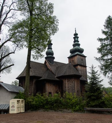 Zdjęcie nr 48: Kościół drewniany o konstrukcji zrębowej, elewacje jednolite, szalowane z listwowaniem, wzmocnione lisicami, wieża o konstrukcji słupowej. Więźba dachowa drewniana, krokwiowa. Budynek na kamiennym podmurowaniu przesłoniętym daszkiem gontowym. Kościół orientowany, jednonawowy z dwiema kaplicami o charakterze transeptu zamkniętymi wielobocznie, na planie krzyża łacińskiego, o węższym prezbiterium zamkniętym trójbocznie. Do wschodniej ściany prezbiterium przylega ogrójec w formie kapliczki, od północy zakrystia na planie prostokąta. Na skrzyżowaniu nawy z południowym ramieniem transeptu kruchta na planie prostokąta. Od zachodu do nawy przylega wieża na planie kwadratu, otoczona sobotami. Prezbiterium i nawa nakryte pozornym sklepieniem tej samej szerokości, obwiedzionym dużym, profilowanym gzymsem podtrzymywanym w nawie przez dwie pary filarów o kanelowanych trzonach, na cokołach. Sklepieniu nawy głównej towarzyszą płaskie i niższe stropy z fasetami w przestrzeniach bocznych. Ściany boczne nawy zwieńczone profilowanym gzymsem. Pomiędzy nawą a prezbiterium otwór tęczowy w formie odcinkowej arkady podtrzymywanej przez filary o kanelowanych trzonach z belką o falistym zarysie. Kaplice boczne otwarte do nawy odcinkowymi arkadami podtrzymywanymi przez filary o kanelowanych trzonach, na cokołach. Parapet chóru muzycznego w zachodniej części nawy, o wybrzuszonej środkowej części, podparty na dwóch filarach o kanelowanych trzonach, na cokołach. Posadzka z płyt ceramicznych. Elewacja frontowa w postaci wieży otoczonej sobotami. Na osi frontowej ściany sobót płytka kruchta zwieńczona gzymsem z przyczółkiem wypełnionym motywem „słoneczka”, przykryta dwuspadowym dachem. Drzwi dwuskrzydłowe, drewniane z okuciami. Korpus wieży podzielony poziomo daszkiem okapowym w dwóch trzecich wysokości, na froncie okno. W dolnej części małe otwory dzwonne z trzech stron, w górnej części duże otwory dzwonne z czterech stron. Wieża zwieńczona hełmem baniastym z latarnią. Ściany sobót szalowane z poziomym listwowaniem, przekryte mansardowym dachem gontowym, na północnej i południowej ścianie dwie pary okien w formie leżącego prostokąta zamkniętego łukiem odcinkowym. W elewacji południowej cztery otwory okienne w formie stojącego prostokąta zamkniętego łukiem odcinkowym, w partii prezbiterium z witrażami. Na elewacji wschodniej ogrójec w formie nadwieszonej kapliczki nakrytej dwuspadowym daszkiem z grupą Ukrzyżowania. W elewacji północnej dwa otwory okienne w formie stojącego prostokąta zamkniętego łukiem odcinkowym. W zakrystii małe kwadratowe okienko. W obu ramionach transeptu analogiczne jak w elewacjach otwory okienne na wschodniej i zachodniej ścianie. Kościół nakryty dachami gontowymi, wyższym nad nawą, niższym nad prezbiterium, dwuspadowym nad nawą oraz wielospadowym nad prezbiterium i ramionami transeptu; nad zakrystią i kruchtą boczną dachy dwuspadowe. W połowie długości dachu, nad nawą, od strony prezbiterium wznosi się ponad kalenicą wieżyczka na sygnaturkę, obita blachą, na ośmiobocznym trzonie, z latarnią zwieńczoną niewielkim baniastym hełmem z latarnią.
