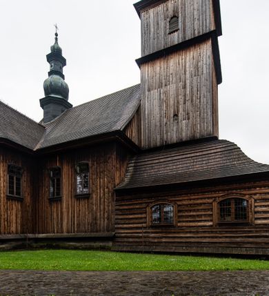 Zdjęcie nr 47: Kościół drewniany o konstrukcji zrębowej, elewacje jednolite, szalowane z listwowaniem, wzmocnione lisicami, wieża o konstrukcji słupowej. Więźba dachowa drewniana, krokwiowa. Budynek na kamiennym podmurowaniu przesłoniętym daszkiem gontowym. Kościół orientowany, jednonawowy z dwiema kaplicami o charakterze transeptu zamkniętymi wielobocznie, na planie krzyża łacińskiego, o węższym prezbiterium zamkniętym trójbocznie. Do wschodniej ściany prezbiterium przylega ogrójec w formie kapliczki, od północy zakrystia na planie prostokąta. Na skrzyżowaniu nawy z południowym ramieniem transeptu kruchta na planie prostokąta. Od zachodu do nawy przylega wieża na planie kwadratu, otoczona sobotami. Prezbiterium i nawa nakryte pozornym sklepieniem tej samej szerokości, obwiedzionym dużym, profilowanym gzymsem podtrzymywanym w nawie przez dwie pary filarów o kanelowanych trzonach, na cokołach. Sklepieniu nawy głównej towarzyszą płaskie i niższe stropy z fasetami w przestrzeniach bocznych. Ściany boczne nawy zwieńczone profilowanym gzymsem. Pomiędzy nawą a prezbiterium otwór tęczowy w formie odcinkowej arkady podtrzymywanej przez filary o kanelowanych trzonach z belką o falistym zarysie. Kaplice boczne otwarte do nawy odcinkowymi arkadami podtrzymywanymi przez filary o kanelowanych trzonach, na cokołach. Parapet chóru muzycznego w zachodniej części nawy, o wybrzuszonej środkowej części, podparty na dwóch filarach o kanelowanych trzonach, na cokołach. Posadzka z płyt ceramicznych. Elewacja frontowa w postaci wieży otoczonej sobotami. Na osi frontowej ściany sobót płytka kruchta zwieńczona gzymsem z przyczółkiem wypełnionym motywem „słoneczka”, przykryta dwuspadowym dachem. Drzwi dwuskrzydłowe, drewniane z okuciami. Korpus wieży podzielony poziomo daszkiem okapowym w dwóch trzecich wysokości, na froncie okno. W dolnej części małe otwory dzwonne z trzech stron, w górnej części duże otwory dzwonne z czterech stron. Wieża zwieńczona hełmem baniastym z latarnią. Ściany sobót szalowane z poziomym listwowaniem, przekryte mansardowym dachem gontowym, na północnej i południowej ścianie dwie pary okien w formie leżącego prostokąta zamkniętego łukiem odcinkowym. W elewacji południowej cztery otwory okienne w formie stojącego prostokąta zamkniętego łukiem odcinkowym, w partii prezbiterium z witrażami. Na elewacji wschodniej ogrójec w formie nadwieszonej kapliczki nakrytej dwuspadowym daszkiem z grupą Ukrzyżowania. W elewacji północnej dwa otwory okienne w formie stojącego prostokąta zamkniętego łukiem odcinkowym. W zakrystii małe kwadratowe okienko. W obu ramionach transeptu analogiczne jak w elewacjach otwory okienne na wschodniej i zachodniej ścianie. Kościół nakryty dachami gontowymi, wyższym nad nawą, niższym nad prezbiterium, dwuspadowym nad nawą oraz wielospadowym nad prezbiterium i ramionami transeptu; nad zakrystią i kruchtą boczną dachy dwuspadowe. W połowie długości dachu, nad nawą, od strony prezbiterium wznosi się ponad kalenicą wieżyczka na sygnaturkę, obita blachą, na ośmiobocznym trzonie, z latarnią zwieńczoną niewielkim baniastym hełmem z latarnią.
