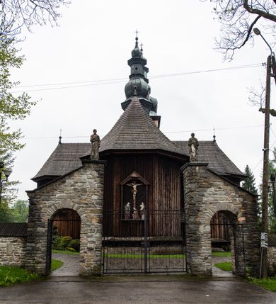 Zdjęcie nr 54: Kościół drewniany o konstrukcji zrębowej, elewacje jednolite, szalowane z listwowaniem, wzmocnione lisicami, wieża o konstrukcji słupowej. Więźba dachowa drewniana, krokwiowa. Budynek na kamiennym podmurowaniu przesłoniętym daszkiem gontowym. Kościół orientowany, jednonawowy z dwiema kaplicami o charakterze transeptu zamkniętymi wielobocznie, na planie krzyża łacińskiego, o węższym prezbiterium zamkniętym trójbocznie. Do wschodniej ściany prezbiterium przylega ogrójec w formie kapliczki, od północy zakrystia na planie prostokąta. Na skrzyżowaniu nawy z południowym ramieniem transeptu kruchta na planie prostokąta. Od zachodu do nawy przylega wieża na planie kwadratu, otoczona sobotami. Prezbiterium i nawa nakryte pozornym sklepieniem tej samej szerokości, obwiedzionym dużym, profilowanym gzymsem podtrzymywanym w nawie przez dwie pary filarów o kanelowanych trzonach, na cokołach. Sklepieniu nawy głównej towarzyszą płaskie i niższe stropy z fasetami w przestrzeniach bocznych. Ściany boczne nawy zwieńczone profilowanym gzymsem. Pomiędzy nawą a prezbiterium otwór tęczowy w formie odcinkowej arkady podtrzymywanej przez filary o kanelowanych trzonach z belką o falistym zarysie. Kaplice boczne otwarte do nawy odcinkowymi arkadami podtrzymywanymi przez filary o kanelowanych trzonach, na cokołach. Parapet chóru muzycznego w zachodniej części nawy, o wybrzuszonej środkowej części, podparty na dwóch filarach o kanelowanych trzonach, na cokołach. Posadzka z płyt ceramicznych. Elewacja frontowa w postaci wieży otoczonej sobotami. Na osi frontowej ściany sobót płytka kruchta zwieńczona gzymsem z przyczółkiem wypełnionym motywem „słoneczka”, przykryta dwuspadowym dachem. Drzwi dwuskrzydłowe, drewniane z okuciami. Korpus wieży podzielony poziomo daszkiem okapowym w dwóch trzecich wysokości, na froncie okno. W dolnej części małe otwory dzwonne z trzech stron, w górnej części duże otwory dzwonne z czterech stron. Wieża zwieńczona hełmem baniastym z latarnią. Ściany sobót szalowane z poziomym listwowaniem, przekryte mansardowym dachem gontowym, na północnej i południowej ścianie dwie pary okien w formie leżącego prostokąta zamkniętego łukiem odcinkowym. W elewacji południowej cztery otwory okienne w formie stojącego prostokąta zamkniętego łukiem odcinkowym, w partii prezbiterium z witrażami. Na elewacji wschodniej ogrójec w formie nadwieszonej kapliczki nakrytej dwuspadowym daszkiem z grupą Ukrzyżowania. W elewacji północnej dwa otwory okienne w formie stojącego prostokąta zamkniętego łukiem odcinkowym. W zakrystii małe kwadratowe okienko. W obu ramionach transeptu analogiczne jak w elewacjach otwory okienne na wschodniej i zachodniej ścianie. Kościół nakryty dachami gontowymi, wyższym nad nawą, niższym nad prezbiterium, dwuspadowym nad nawą oraz wielospadowym nad prezbiterium i ramionami transeptu; nad zakrystią i kruchtą boczną dachy dwuspadowe. W połowie długości dachu, nad nawą, od strony prezbiterium wznosi się ponad kalenicą wieżyczka na sygnaturkę, obita blachą, na ośmiobocznym trzonie, z latarnią zwieńczoną niewielkim baniastym hełmem z latarnią.
