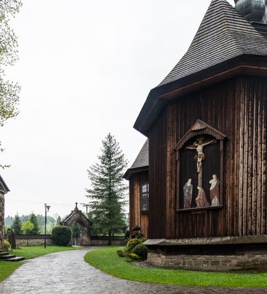 Zdjęcie nr 56: Kościół drewniany o konstrukcji zrębowej, elewacje jednolite, szalowane z listwowaniem, wzmocnione lisicami, wieża o konstrukcji słupowej. Więźba dachowa drewniana, krokwiowa. Budynek na kamiennym podmurowaniu przesłoniętym daszkiem gontowym. Kościół orientowany, jednonawowy z dwiema kaplicami o charakterze transeptu zamkniętymi wielobocznie, na planie krzyża łacińskiego, o węższym prezbiterium zamkniętym trójbocznie. Do wschodniej ściany prezbiterium przylega ogrójec w formie kapliczki, od północy zakrystia na planie prostokąta. Na skrzyżowaniu nawy z południowym ramieniem transeptu kruchta na planie prostokąta. Od zachodu do nawy przylega wieża na planie kwadratu, otoczona sobotami. Prezbiterium i nawa nakryte pozornym sklepieniem tej samej szerokości, obwiedzionym dużym, profilowanym gzymsem podtrzymywanym w nawie przez dwie pary filarów o kanelowanych trzonach, na cokołach. Sklepieniu nawy głównej towarzyszą płaskie i niższe stropy z fasetami w przestrzeniach bocznych. Ściany boczne nawy zwieńczone profilowanym gzymsem. Pomiędzy nawą a prezbiterium otwór tęczowy w formie odcinkowej arkady podtrzymywanej przez filary o kanelowanych trzonach z belką o falistym zarysie. Kaplice boczne otwarte do nawy odcinkowymi arkadami podtrzymywanymi przez filary o kanelowanych trzonach, na cokołach. Parapet chóru muzycznego w zachodniej części nawy, o wybrzuszonej środkowej części, podparty na dwóch filarach o kanelowanych trzonach, na cokołach. Posadzka z płyt ceramicznych. Elewacja frontowa w postaci wieży otoczonej sobotami. Na osi frontowej ściany sobót płytka kruchta zwieńczona gzymsem z przyczółkiem wypełnionym motywem „słoneczka”, przykryta dwuspadowym dachem. Drzwi dwuskrzydłowe, drewniane z okuciami. Korpus wieży podzielony poziomo daszkiem okapowym w dwóch trzecich wysokości, na froncie okno. W dolnej części małe otwory dzwonne z trzech stron, w górnej części duże otwory dzwonne z czterech stron. Wieża zwieńczona hełmem baniastym z latarnią. Ściany sobót szalowane z poziomym listwowaniem, przekryte mansardowym dachem gontowym, na północnej i południowej ścianie dwie pary okien w formie leżącego prostokąta zamkniętego łukiem odcinkowym. W elewacji południowej cztery otwory okienne w formie stojącego prostokąta zamkniętego łukiem odcinkowym, w partii prezbiterium z witrażami. Na elewacji wschodniej ogrójec w formie nadwieszonej kapliczki nakrytej dwuspadowym daszkiem z grupą Ukrzyżowania. W elewacji północnej dwa otwory okienne w formie stojącego prostokąta zamkniętego łukiem odcinkowym. W zakrystii małe kwadratowe okienko. W obu ramionach transeptu analogiczne jak w elewacjach otwory okienne na wschodniej i zachodniej ścianie. Kościół nakryty dachami gontowymi, wyższym nad nawą, niższym nad prezbiterium, dwuspadowym nad nawą oraz wielospadowym nad prezbiterium i ramionami transeptu; nad zakrystią i kruchtą boczną dachy dwuspadowe. W połowie długości dachu, nad nawą, od strony prezbiterium wznosi się ponad kalenicą wieżyczka na sygnaturkę, obita blachą, na ośmiobocznym trzonie, z latarnią zwieńczoną niewielkim baniastym hełmem z latarnią.
