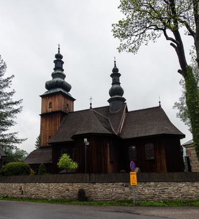 Zdjęcie nr 23: Kościół drewniany o konstrukcji zrębowej, elewacje jednolite, szalowane z listwowaniem, wzmocnione lisicami, wieża o konstrukcji słupowej. Więźba dachowa drewniana, krokwiowa. Budynek na kamiennym podmurowaniu przesłoniętym daszkiem gontowym. Kościół orientowany, jednonawowy z dwiema kaplicami o charakterze transeptu zamkniętymi wielobocznie, na planie krzyża łacińskiego, o węższym prezbiterium zamkniętym trójbocznie. Do wschodniej ściany prezbiterium przylega ogrójec w formie kapliczki, od północy zakrystia na planie prostokąta. Na skrzyżowaniu nawy z południowym ramieniem transeptu kruchta na planie prostokąta. Od zachodu do nawy przylega wieża na planie kwadratu, otoczona sobotami. Prezbiterium i nawa nakryte pozornym sklepieniem tej samej szerokości, obwiedzionym dużym, profilowanym gzymsem podtrzymywanym w nawie przez dwie pary filarów o kanelowanych trzonach, na cokołach. Sklepieniu nawy głównej towarzyszą płaskie i niższe stropy z fasetami w przestrzeniach bocznych. Ściany boczne nawy zwieńczone profilowanym gzymsem. Pomiędzy nawą a prezbiterium otwór tęczowy w formie odcinkowej arkady podtrzymywanej przez filary o kanelowanych trzonach z belką o falistym zarysie. Kaplice boczne otwarte do nawy odcinkowymi arkadami podtrzymywanymi przez filary o kanelowanych trzonach, na cokołach. Parapet chóru muzycznego w zachodniej części nawy, o wybrzuszonej środkowej części, podparty na dwóch filarach o kanelowanych trzonach, na cokołach. Posadzka z płyt ceramicznych. Elewacja frontowa w postaci wieży otoczonej sobotami. Na osi frontowej ściany sobót płytka kruchta zwieńczona gzymsem z przyczółkiem wypełnionym motywem „słoneczka”, przykryta dwuspadowym dachem. Drzwi dwuskrzydłowe, drewniane z okuciami. Korpus wieży podzielony poziomo daszkiem okapowym w dwóch trzecich wysokości, na froncie okno. W dolnej części małe otwory dzwonne z trzech stron, w górnej części duże otwory dzwonne z czterech stron. Wieża zwieńczona hełmem baniastym z latarnią. Ściany sobót szalowane z poziomym listwowaniem, przekryte mansardowym dachem gontowym, na północnej i południowej ścianie dwie pary okien w formie leżącego prostokąta zamkniętego łukiem odcinkowym. W elewacji południowej cztery otwory okienne w formie stojącego prostokąta zamkniętego łukiem odcinkowym, w partii prezbiterium z witrażami. Na elewacji wschodniej ogrójec w formie nadwieszonej kapliczki nakrytej dwuspadowym daszkiem z grupą Ukrzyżowania. W elewacji północnej dwa otwory okienne w formie stojącego prostokąta zamkniętego łukiem odcinkowym. W zakrystii małe kwadratowe okienko. W obu ramionach transeptu analogiczne jak w elewacjach otwory okienne na wschodniej i zachodniej ścianie. Kościół nakryty dachami gontowymi, wyższym nad nawą, niższym nad prezbiterium, dwuspadowym nad nawą oraz wielospadowym nad prezbiterium i ramionami transeptu; nad zakrystią i kruchtą boczną dachy dwuspadowe. W połowie długości dachu, nad nawą, od strony prezbiterium wznosi się ponad kalenicą wieżyczka na sygnaturkę, obita blachą, na ośmiobocznym trzonie, z latarnią zwieńczoną niewielkim baniastym hełmem z latarnią.
