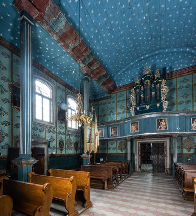 Zdjęcie nr 8: Kościół drewniany o konstrukcji zrębowej, elewacje jednolite, szalowane z listwowaniem, wzmocnione lisicami, wieża o konstrukcji słupowej. Więźba dachowa drewniana, krokwiowa. Budynek na kamiennym podmurowaniu przesłoniętym daszkiem gontowym. Kościół orientowany, jednonawowy z dwiema kaplicami o charakterze transeptu zamkniętymi wielobocznie, na planie krzyża łacińskiego, o węższym prezbiterium zamkniętym trójbocznie. Do wschodniej ściany prezbiterium przylega ogrójec w formie kapliczki, od północy zakrystia na planie prostokąta. Na skrzyżowaniu nawy z południowym ramieniem transeptu kruchta na planie prostokąta. Od zachodu do nawy przylega wieża na planie kwadratu, otoczona sobotami. Prezbiterium i nawa nakryte pozornym sklepieniem tej samej szerokości, obwiedzionym dużym, profilowanym gzymsem podtrzymywanym w nawie przez dwie pary filarów o kanelowanych trzonach, na cokołach. Sklepieniu nawy głównej towarzyszą płaskie i niższe stropy z fasetami w przestrzeniach bocznych. Ściany boczne nawy zwieńczone profilowanym gzymsem. Pomiędzy nawą a prezbiterium otwór tęczowy w formie odcinkowej arkady podtrzymywanej przez filary o kanelowanych trzonach z belką o falistym zarysie. Kaplice boczne otwarte do nawy odcinkowymi arkadami podtrzymywanymi przez filary o kanelowanych trzonach, na cokołach. Parapet chóru muzycznego w zachodniej części nawy, o wybrzuszonej środkowej części, podparty na dwóch filarach o kanelowanych trzonach, na cokołach. Posadzka z płyt ceramicznych. Elewacja frontowa w postaci wieży otoczonej sobotami. Na osi frontowej ściany sobót płytka kruchta zwieńczona gzymsem z przyczółkiem wypełnionym motywem „słoneczka”, przykryta dwuspadowym dachem. Drzwi dwuskrzydłowe, drewniane z okuciami. Korpus wieży podzielony poziomo daszkiem okapowym w dwóch trzecich wysokości, na froncie okno. W dolnej części małe otwory dzwonne z trzech stron, w górnej części duże otwory dzwonne z czterech stron. Wieża zwieńczona hełmem baniastym z latarnią. Ściany sobót szalowane z poziomym listwowaniem, przekryte mansardowym dachem gontowym, na północnej i południowej ścianie dwie pary okien w formie leżącego prostokąta zamkniętego łukiem odcinkowym. W elewacji południowej cztery otwory okienne w formie stojącego prostokąta zamkniętego łukiem odcinkowym, w partii prezbiterium z witrażami. Na elewacji wschodniej ogrójec w formie nadwieszonej kapliczki nakrytej dwuspadowym daszkiem z grupą Ukrzyżowania. W elewacji północnej dwa otwory okienne w formie stojącego prostokąta zamkniętego łukiem odcinkowym. W zakrystii małe kwadratowe okienko. W obu ramionach transeptu analogiczne jak w elewacjach otwory okienne na wschodniej i zachodniej ścianie. Kościół nakryty dachami gontowymi, wyższym nad nawą, niższym nad prezbiterium, dwuspadowym nad nawą oraz wielospadowym nad prezbiterium i ramionami transeptu; nad zakrystią i kruchtą boczną dachy dwuspadowe. W połowie długości dachu, nad nawą, od strony prezbiterium wznosi się ponad kalenicą wieżyczka na sygnaturkę, obita blachą, na ośmiobocznym trzonie, z latarnią zwieńczoną niewielkim baniastym hełmem z latarnią.
