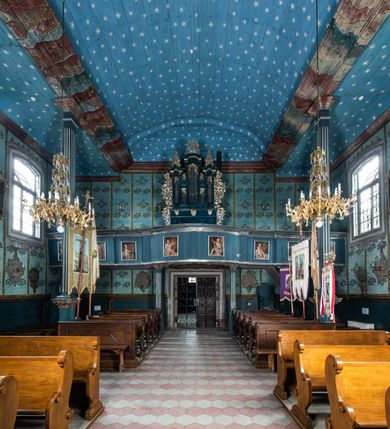 Zdjęcie nr 5: Kościół drewniany o konstrukcji zrębowej, elewacje jednolite, szalowane z listwowaniem, wzmocnione lisicami, wieża o konstrukcji słupowej. Więźba dachowa drewniana, krokwiowa. Budynek na kamiennym podmurowaniu przesłoniętym daszkiem gontowym. Kościół orientowany, jednonawowy z dwiema kaplicami o charakterze transeptu zamkniętymi wielobocznie, na planie krzyża łacińskiego, o węższym prezbiterium zamkniętym trójbocznie. Do wschodniej ściany prezbiterium przylega ogrójec w formie kapliczki, od północy zakrystia na planie prostokąta. Na skrzyżowaniu nawy z południowym ramieniem transeptu kruchta na planie prostokąta. Od zachodu do nawy przylega wieża na planie kwadratu, otoczona sobotami. Prezbiterium i nawa nakryte pozornym sklepieniem tej samej szerokości, obwiedzionym dużym, profilowanym gzymsem podtrzymywanym w nawie przez dwie pary filarów o kanelowanych trzonach, na cokołach. Sklepieniu nawy głównej towarzyszą płaskie i niższe stropy z fasetami w przestrzeniach bocznych. Ściany boczne nawy zwieńczone profilowanym gzymsem. Pomiędzy nawą a prezbiterium otwór tęczowy w formie odcinkowej arkady podtrzymywanej przez filary o kanelowanych trzonach z belką o falistym zarysie. Kaplice boczne otwarte do nawy odcinkowymi arkadami podtrzymywanymi przez filary o kanelowanych trzonach, na cokołach. Parapet chóru muzycznego w zachodniej części nawy, o wybrzuszonej środkowej części, podparty na dwóch filarach o kanelowanych trzonach, na cokołach. Posadzka z płyt ceramicznych. Elewacja frontowa w postaci wieży otoczonej sobotami. Na osi frontowej ściany sobót płytka kruchta zwieńczona gzymsem z przyczółkiem wypełnionym motywem „słoneczka”, przykryta dwuspadowym dachem. Drzwi dwuskrzydłowe, drewniane z okuciami. Korpus wieży podzielony poziomo daszkiem okapowym w dwóch trzecich wysokości, na froncie okno. W dolnej części małe otwory dzwonne z trzech stron, w górnej części duże otwory dzwonne z czterech stron. Wieża zwieńczona hełmem baniastym z latarnią. Ściany sobót szalowane z poziomym listwowaniem, przekryte mansardowym dachem gontowym, na północnej i południowej ścianie dwie pary okien w formie leżącego prostokąta zamkniętego łukiem odcinkowym. W elewacji południowej cztery otwory okienne w formie stojącego prostokąta zamkniętego łukiem odcinkowym, w partii prezbiterium z witrażami. Na elewacji wschodniej ogrójec w formie nadwieszonej kapliczki nakrytej dwuspadowym daszkiem z grupą Ukrzyżowania. W elewacji północnej dwa otwory okienne w formie stojącego prostokąta zamkniętego łukiem odcinkowym. W zakrystii małe kwadratowe okienko. W obu ramionach transeptu analogiczne jak w elewacjach otwory okienne na wschodniej i zachodniej ścianie. Kościół nakryty dachami gontowymi, wyższym nad nawą, niższym nad prezbiterium, dwuspadowym nad nawą oraz wielospadowym nad prezbiterium i ramionami transeptu; nad zakrystią i kruchtą boczną dachy dwuspadowe. W połowie długości dachu, nad nawą, od strony prezbiterium wznosi się ponad kalenicą wieżyczka na sygnaturkę, obita blachą, na ośmiobocznym trzonie, z latarnią zwieńczoną niewielkim baniastym hełmem z latarnią.
