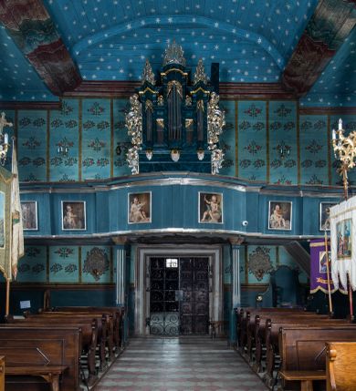 Zdjęcie nr 6: Kościół drewniany o konstrukcji zrębowej, elewacje jednolite, szalowane z listwowaniem, wzmocnione lisicami, wieża o konstrukcji słupowej. Więźba dachowa drewniana, krokwiowa. Budynek na kamiennym podmurowaniu przesłoniętym daszkiem gontowym. Kościół orientowany, jednonawowy z dwiema kaplicami o charakterze transeptu zamkniętymi wielobocznie, na planie krzyża łacińskiego, o węższym prezbiterium zamkniętym trójbocznie. Do wschodniej ściany prezbiterium przylega ogrójec w formie kapliczki, od północy zakrystia na planie prostokąta. Na skrzyżowaniu nawy z południowym ramieniem transeptu kruchta na planie prostokąta. Od zachodu do nawy przylega wieża na planie kwadratu, otoczona sobotami. Prezbiterium i nawa nakryte pozornym sklepieniem tej samej szerokości, obwiedzionym dużym, profilowanym gzymsem podtrzymywanym w nawie przez dwie pary filarów o kanelowanych trzonach, na cokołach. Sklepieniu nawy głównej towarzyszą płaskie i niższe stropy z fasetami w przestrzeniach bocznych. Ściany boczne nawy zwieńczone profilowanym gzymsem. Pomiędzy nawą a prezbiterium otwór tęczowy w formie odcinkowej arkady podtrzymywanej przez filary o kanelowanych trzonach z belką o falistym zarysie. Kaplice boczne otwarte do nawy odcinkowymi arkadami podtrzymywanymi przez filary o kanelowanych trzonach, na cokołach. Parapet chóru muzycznego w zachodniej części nawy, o wybrzuszonej środkowej części, podparty na dwóch filarach o kanelowanych trzonach, na cokołach. Posadzka z płyt ceramicznych. Elewacja frontowa w postaci wieży otoczonej sobotami. Na osi frontowej ściany sobót płytka kruchta zwieńczona gzymsem z przyczółkiem wypełnionym motywem „słoneczka”, przykryta dwuspadowym dachem. Drzwi dwuskrzydłowe, drewniane z okuciami. Korpus wieży podzielony poziomo daszkiem okapowym w dwóch trzecich wysokości, na froncie okno. W dolnej części małe otwory dzwonne z trzech stron, w górnej części duże otwory dzwonne z czterech stron. Wieża zwieńczona hełmem baniastym z latarnią. Ściany sobót szalowane z poziomym listwowaniem, przekryte mansardowym dachem gontowym, na północnej i południowej ścianie dwie pary okien w formie leżącego prostokąta zamkniętego łukiem odcinkowym. W elewacji południowej cztery otwory okienne w formie stojącego prostokąta zamkniętego łukiem odcinkowym, w partii prezbiterium z witrażami. Na elewacji wschodniej ogrójec w formie nadwieszonej kapliczki nakrytej dwuspadowym daszkiem z grupą Ukrzyżowania. W elewacji północnej dwa otwory okienne w formie stojącego prostokąta zamkniętego łukiem odcinkowym. W zakrystii małe kwadratowe okienko. W obu ramionach transeptu analogiczne jak w elewacjach otwory okienne na wschodniej i zachodniej ścianie. Kościół nakryty dachami gontowymi, wyższym nad nawą, niższym nad prezbiterium, dwuspadowym nad nawą oraz wielospadowym nad prezbiterium i ramionami transeptu; nad zakrystią i kruchtą boczną dachy dwuspadowe. W połowie długości dachu, nad nawą, od strony prezbiterium wznosi się ponad kalenicą wieżyczka na sygnaturkę, obita blachą, na ośmiobocznym trzonie, z latarnią zwieńczoną niewielkim baniastym hełmem z latarnią.
