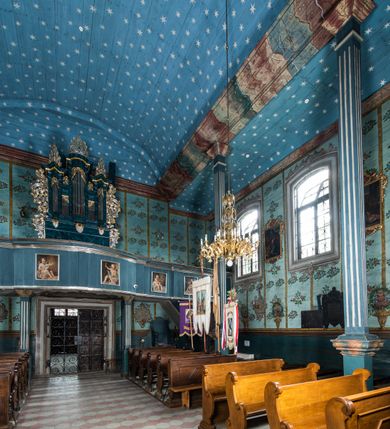 Zdjęcie nr 7: Kościół drewniany o konstrukcji zrębowej, elewacje jednolite, szalowane z listwowaniem, wzmocnione lisicami, wieża o konstrukcji słupowej. Więźba dachowa drewniana, krokwiowa. Budynek na kamiennym podmurowaniu przesłoniętym daszkiem gontowym. Kościół orientowany, jednonawowy z dwiema kaplicami o charakterze transeptu zamkniętymi wielobocznie, na planie krzyża łacińskiego, o węższym prezbiterium zamkniętym trójbocznie. Do wschodniej ściany prezbiterium przylega ogrójec w formie kapliczki, od północy zakrystia na planie prostokąta. Na skrzyżowaniu nawy z południowym ramieniem transeptu kruchta na planie prostokąta. Od zachodu do nawy przylega wieża na planie kwadratu, otoczona sobotami. Prezbiterium i nawa nakryte pozornym sklepieniem tej samej szerokości, obwiedzionym dużym, profilowanym gzymsem podtrzymywanym w nawie przez dwie pary filarów o kanelowanych trzonach, na cokołach. Sklepieniu nawy głównej towarzyszą płaskie i niższe stropy z fasetami w przestrzeniach bocznych. Ściany boczne nawy zwieńczone profilowanym gzymsem. Pomiędzy nawą a prezbiterium otwór tęczowy w formie odcinkowej arkady podtrzymywanej przez filary o kanelowanych trzonach z belką o falistym zarysie. Kaplice boczne otwarte do nawy odcinkowymi arkadami podtrzymywanymi przez filary o kanelowanych trzonach, na cokołach. Parapet chóru muzycznego w zachodniej części nawy, o wybrzuszonej środkowej części, podparty na dwóch filarach o kanelowanych trzonach, na cokołach. Posadzka z płyt ceramicznych. Elewacja frontowa w postaci wieży otoczonej sobotami. Na osi frontowej ściany sobót płytka kruchta zwieńczona gzymsem z przyczółkiem wypełnionym motywem „słoneczka”, przykryta dwuspadowym dachem. Drzwi dwuskrzydłowe, drewniane z okuciami. Korpus wieży podzielony poziomo daszkiem okapowym w dwóch trzecich wysokości, na froncie okno. W dolnej części małe otwory dzwonne z trzech stron, w górnej części duże otwory dzwonne z czterech stron. Wieża zwieńczona hełmem baniastym z latarnią. Ściany sobót szalowane z poziomym listwowaniem, przekryte mansardowym dachem gontowym, na północnej i południowej ścianie dwie pary okien w formie leżącego prostokąta zamkniętego łukiem odcinkowym. W elewacji południowej cztery otwory okienne w formie stojącego prostokąta zamkniętego łukiem odcinkowym, w partii prezbiterium z witrażami. Na elewacji wschodniej ogrójec w formie nadwieszonej kapliczki nakrytej dwuspadowym daszkiem z grupą Ukrzyżowania. W elewacji północnej dwa otwory okienne w formie stojącego prostokąta zamkniętego łukiem odcinkowym. W zakrystii małe kwadratowe okienko. W obu ramionach transeptu analogiczne jak w elewacjach otwory okienne na wschodniej i zachodniej ścianie. Kościół nakryty dachami gontowymi, wyższym nad nawą, niższym nad prezbiterium, dwuspadowym nad nawą oraz wielospadowym nad prezbiterium i ramionami transeptu; nad zakrystią i kruchtą boczną dachy dwuspadowe. W połowie długości dachu, nad nawą, od strony prezbiterium wznosi się ponad kalenicą wieżyczka na sygnaturkę, obita blachą, na ośmiobocznym trzonie, z latarnią zwieńczoną niewielkim baniastym hełmem z latarnią.
