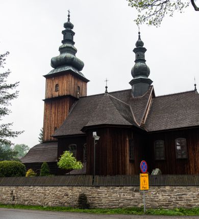 Zdjęcie nr 24: Kościół drewniany o konstrukcji zrębowej, elewacje jednolite, szalowane z listwowaniem, wzmocnione lisicami, wieża o konstrukcji słupowej. Więźba dachowa drewniana, krokwiowa. Budynek na kamiennym podmurowaniu przesłoniętym daszkiem gontowym. Kościół orientowany, jednonawowy z dwiema kaplicami o charakterze transeptu zamkniętymi wielobocznie, na planie krzyża łacińskiego, o węższym prezbiterium zamkniętym trójbocznie. Do wschodniej ściany prezbiterium przylega ogrójec w formie kapliczki, od północy zakrystia na planie prostokąta. Na skrzyżowaniu nawy z południowym ramieniem transeptu kruchta na planie prostokąta. Od zachodu do nawy przylega wieża na planie kwadratu, otoczona sobotami. Prezbiterium i nawa nakryte pozornym sklepieniem tej samej szerokości, obwiedzionym dużym, profilowanym gzymsem podtrzymywanym w nawie przez dwie pary filarów o kanelowanych trzonach, na cokołach. Sklepieniu nawy głównej towarzyszą płaskie i niższe stropy z fasetami w przestrzeniach bocznych. Ściany boczne nawy zwieńczone profilowanym gzymsem. Pomiędzy nawą a prezbiterium otwór tęczowy w formie odcinkowej arkady podtrzymywanej przez filary o kanelowanych trzonach z belką o falistym zarysie. Kaplice boczne otwarte do nawy odcinkowymi arkadami podtrzymywanymi przez filary o kanelowanych trzonach, na cokołach. Parapet chóru muzycznego w zachodniej części nawy, o wybrzuszonej środkowej części, podparty na dwóch filarach o kanelowanych trzonach, na cokołach. Posadzka z płyt ceramicznych. Elewacja frontowa w postaci wieży otoczonej sobotami. Na osi frontowej ściany sobót płytka kruchta zwieńczona gzymsem z przyczółkiem wypełnionym motywem „słoneczka”, przykryta dwuspadowym dachem. Drzwi dwuskrzydłowe, drewniane z okuciami. Korpus wieży podzielony poziomo daszkiem okapowym w dwóch trzecich wysokości, na froncie okno. W dolnej części małe otwory dzwonne z trzech stron, w górnej części duże otwory dzwonne z czterech stron. Wieża zwieńczona hełmem baniastym z latarnią. Ściany sobót szalowane z poziomym listwowaniem, przekryte mansardowym dachem gontowym, na północnej i południowej ścianie dwie pary okien w formie leżącego prostokąta zamkniętego łukiem odcinkowym. W elewacji południowej cztery otwory okienne w formie stojącego prostokąta zamkniętego łukiem odcinkowym, w partii prezbiterium z witrażami. Na elewacji wschodniej ogrójec w formie nadwieszonej kapliczki nakrytej dwuspadowym daszkiem z grupą Ukrzyżowania. W elewacji północnej dwa otwory okienne w formie stojącego prostokąta zamkniętego łukiem odcinkowym. W zakrystii małe kwadratowe okienko. W obu ramionach transeptu analogiczne jak w elewacjach otwory okienne na wschodniej i zachodniej ścianie. Kościół nakryty dachami gontowymi, wyższym nad nawą, niższym nad prezbiterium, dwuspadowym nad nawą oraz wielospadowym nad prezbiterium i ramionami transeptu; nad zakrystią i kruchtą boczną dachy dwuspadowe. W połowie długości dachu, nad nawą, od strony prezbiterium wznosi się ponad kalenicą wieżyczka na sygnaturkę, obita blachą, na ośmiobocznym trzonie, z latarnią zwieńczoną niewielkim baniastym hełmem z latarnią.
