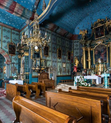 Zdjęcie nr 3: Kościół drewniany o konstrukcji zrębowej, elewacje jednolite, szalowane z listwowaniem, wzmocnione lisicami, wieża o konstrukcji słupowej. Więźba dachowa drewniana, krokwiowa. Budynek na kamiennym podmurowaniu przesłoniętym daszkiem gontowym. Kościół orientowany, jednonawowy z dwiema kaplicami o charakterze transeptu zamkniętymi wielobocznie, na planie krzyża łacińskiego, o węższym prezbiterium zamkniętym trójbocznie. Do wschodniej ściany prezbiterium przylega ogrójec w formie kapliczki, od północy zakrystia na planie prostokąta. Na skrzyżowaniu nawy z południowym ramieniem transeptu kruchta na planie prostokąta. Od zachodu do nawy przylega wieża na planie kwadratu, otoczona sobotami. Prezbiterium i nawa nakryte pozornym sklepieniem tej samej szerokości, obwiedzionym dużym, profilowanym gzymsem podtrzymywanym w nawie przez dwie pary filarów o kanelowanych trzonach, na cokołach. Sklepieniu nawy głównej towarzyszą płaskie i niższe stropy z fasetami w przestrzeniach bocznych. Ściany boczne nawy zwieńczone profilowanym gzymsem. Pomiędzy nawą a prezbiterium otwór tęczowy w formie odcinkowej arkady podtrzymywanej przez filary o kanelowanych trzonach z belką o falistym zarysie. Kaplice boczne otwarte do nawy odcinkowymi arkadami podtrzymywanymi przez filary o kanelowanych trzonach, na cokołach. Parapet chóru muzycznego w zachodniej części nawy, o wybrzuszonej środkowej części, podparty na dwóch filarach o kanelowanych trzonach, na cokołach. Posadzka z płyt ceramicznych. Elewacja frontowa w postaci wieży otoczonej sobotami. Na osi frontowej ściany sobót płytka kruchta zwieńczona gzymsem z przyczółkiem wypełnionym motywem „słoneczka”, przykryta dwuspadowym dachem. Drzwi dwuskrzydłowe, drewniane z okuciami. Korpus wieży podzielony poziomo daszkiem okapowym w dwóch trzecich wysokości, na froncie okno. W dolnej części małe otwory dzwonne z trzech stron, w górnej części duże otwory dzwonne z czterech stron. Wieża zwieńczona hełmem baniastym z latarnią. Ściany sobót szalowane z poziomym listwowaniem, przekryte mansardowym dachem gontowym, na północnej i południowej ścianie dwie pary okien w formie leżącego prostokąta zamkniętego łukiem odcinkowym. W elewacji południowej cztery otwory okienne w formie stojącego prostokąta zamkniętego łukiem odcinkowym, w partii prezbiterium z witrażami. Na elewacji wschodniej ogrójec w formie nadwieszonej kapliczki nakrytej dwuspadowym daszkiem z grupą Ukrzyżowania. W elewacji północnej dwa otwory okienne w formie stojącego prostokąta zamkniętego łukiem odcinkowym. W zakrystii małe kwadratowe okienko. W obu ramionach transeptu analogiczne jak w elewacjach otwory okienne na wschodniej i zachodniej ścianie. Kościół nakryty dachami gontowymi, wyższym nad nawą, niższym nad prezbiterium, dwuspadowym nad nawą oraz wielospadowym nad prezbiterium i ramionami transeptu; nad zakrystią i kruchtą boczną dachy dwuspadowe. W połowie długości dachu, nad nawą, od strony prezbiterium wznosi się ponad kalenicą wieżyczka na sygnaturkę, obita blachą, na ośmiobocznym trzonie, z latarnią zwieńczoną niewielkim baniastym hełmem z latarnią.
