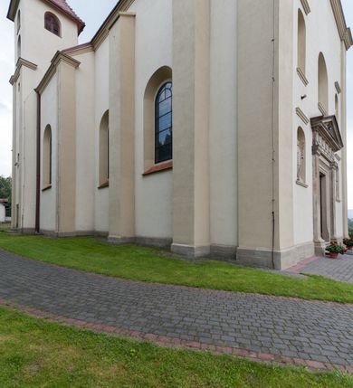 Zdjęcie nr 3: Kościół jest jednonawowy, trójprzęsłowy, z płytkimi kaplicami tworzącymi pseudotransept i jednoprzęsłowym prezbiterium zakończonym apsydą. Do prezbiterium przylegają dwa aneksy mieszące w przyziemiu zakrystię i skarbczyk, a na piętrze empory otwarte na prezbiterium. Do nawy od południa dostawiona jest kruchta. W naroża między kaplicami a nawą wstawiono dwie wieże-dzwonnice z klatkami schodowymi prowadzącymi na empory. Nawa jest rozczłonkowana płaskimi, szerokimi pilastrami toskańskimi, a nakryta sklepieniem kolebkowym z lunetami; prezbiterium przykryte sklepieniem kolebkowym z żebrami spływającymi na wsporniki, apsyda sklepieniem parasolowatym.  Prezbiterium ujęte dwiema głębokimi, półkoliście zamkniętymi wnękami, nad którymi parapety empor z ślepymi arkadkami z balasów jenolalkowych i łuków półkolistych. Wnętrze oświetlają wysokie i szerokie okna zamknięte półkoliście. W aneksach przy prezbiterium i w emporach znajdują się okna prostokątne. Fasada dwustrefowa, trójosiowa, ale osie i strefy wyznaczone jedynie przez okna, drzwi i nisze, zamknięta trójkątnie.    
Portal prostokątnych drzwi utworzony z pary żłobkowanych, toskańskich pilastrów, podtrzymujących belkowanie z trójkątnym przyczółkiem. Fryz belkowania ozdobiony tryglifami z łezkami i metopami w kształcie rozet i talerzy, a przyczółek kostkowaniem. Nad portalem okno w głębokim rozglifieniu, w kształcie prostokąta zamkniętego półkoliście. Po bokach portalu i okna głębokie nisze ujęte od dołu i góry odcinkami gzymsu. Zewnętrzne elewacje ujęte częściowo szkarpami, częściowo lizenami. W wieżach wejścia do kościoła i na klatki schodowe oraz niewielkie okienka w klatce schodowej i prześwity zamknięte półkoliście w dzwonnicach. Dachy dwuspadowe, na wieżach czterospadowe, nad nawą, wieżami i aneksami przy prezbiterium kryte dachówką, nad prezbiterium blachą. Na dachu wieżyczka na sygnaturkę. 
