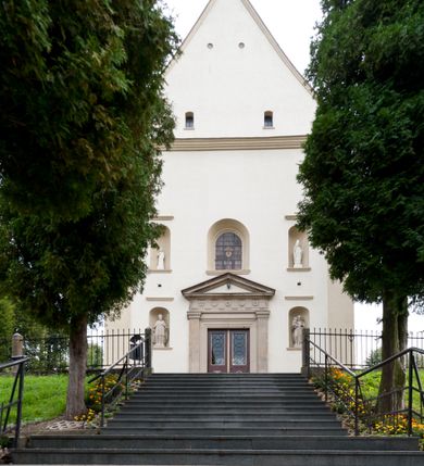 Zdjęcie nr 26: Kościół jest jednonawowy, trójprzęsłowy, z płytkimi kaplicami tworzącymi pseudotransept i jednoprzęsłowym prezbiterium zakończonym apsydą. Do prezbiterium przylegają dwa aneksy mieszące w przyziemiu zakrystię i skarbczyk, a na piętrze empory otwarte na prezbiterium. Do nawy od południa dostawiona jest kruchta. W naroża między kaplicami a nawą wstawiono dwie wieże-dzwonnice z klatkami schodowymi prowadzącymi na empory. Nawa jest rozczłonkowana płaskimi, szerokimi pilastrami toskańskimi, a nakryta sklepieniem kolebkowym z lunetami; prezbiterium przykryte sklepieniem kolebkowym z żebrami spływającymi na wsporniki, apsyda sklepieniem parasolowatym.  Prezbiterium ujęte dwiema głębokimi, półkoliście zamkniętymi wnękami, nad którymi parapety empor z ślepymi arkadkami z balasów jenolalkowych i łuków półkolistych. Wnętrze oświetlają wysokie i szerokie okna zamknięte półkoliście. W aneksach przy prezbiterium i w emporach znajdują się okna prostokątne. Fasada dwustrefowa, trójosiowa, ale osie i strefy wyznaczone jedynie przez okna, drzwi i nisze, zamknięta trójkątnie.    
Portal prostokątnych drzwi utworzony z pary żłobkowanych, toskańskich pilastrów, podtrzymujących belkowanie z trójkątnym przyczółkiem. Fryz belkowania ozdobiony tryglifami z łezkami i metopami w kształcie rozet i talerzy, a przyczółek kostkowaniem. Nad portalem okno w głębokim rozglifieniu, w kształcie prostokąta zamkniętego półkoliście. Po bokach portalu i okna głębokie nisze ujęte od dołu i góry odcinkami gzymsu. Zewnętrzne elewacje ujęte częściowo szkarpami, częściowo lizenami. W wieżach wejścia do kościoła i na klatki schodowe oraz niewielkie okienka w klatce schodowej i prześwity zamknięte półkoliście w dzwonnicach. Dachy dwuspadowe, na wieżach czterospadowe, nad nawą, wieżami i aneksami przy prezbiterium kryte dachówką, nad prezbiterium blachą. Na dachu wieżyczka na sygnaturkę. 

