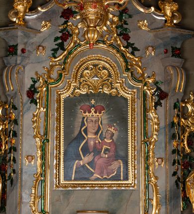 Zdjęcie nr 2: Obraz w kształcie stojącego prostokąta zamkniętego półkolistym łukiem nadwieszonym, uskokowym. Madonna w typie Hodegetrii została przedstawiona frontalnie, w półpostaci. Głowę ma lekko zwróconą w lewo, twarz owalną, o prostych rysach, jasnej karnacji, długim, wąskim nosie, dużych oczach w jasnej oprawie i drobnych ustach. Na prawym policzku dwa równoległe, ukośnie biegnące nacięcia. Maria ubrana jest w jasnoczerwoną suknię ze złoconym galonem pod szyją oraz dekorowany wzorami floralnymi ciemnoniebieski płaszcz ze złotą lamówką osłaniający głowę, ręce i korpus. Na szyi ma zawieszony podwójny sznur pereł ze złotym krzyżykiem, na głowie złotą koronę w typie otwartym, wysadzaną kamieniami szlachetnymi, z czerwonym wypełnieniem. Prawą ręką wskazuje na trzymane na lewej ręce siedzące Dzieciątko. Jezus jest zwrócony w trzech czwartych w prawo. Głowę ma lekko odchyloną na lewy bark, twarz owalną, o jasnej cerze i delikatnych rysach. Długie, brązowe, falowane włosy opadają mu na ramiona i plecy. Jest ubrany w czerwoną tunikę z wąskimi rękawami i złotymi obszyciami. Na głowie ma koronę w typie otwartym, wysadzaną szlachetnymi kamieniami z czerwonym wypełnieniem. Lewą ręką podtrzymuje księgę w oprawie ze złotym napisem IHS i zapięciami, prawą unosi w geście błogosławieństwa. Wokół głów postaci promieniste nimby i gwiazdki. Tło jednolite, szare. W górnej partii obrazu płaskorzeźbiony, ażurowy ornament stylizowanej wici roślinnej z okiem opatrzności Bożej w promienistej glorii pośrodku. Kolorystyka intensywna, mocne kontrasty kolorystyczne. Obraz ujęty złoconą, profilowaną ramą. 