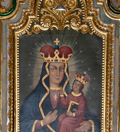 Zdjęcie nr 3: Obraz w kształcie stojącego prostokąta zamkniętego półkolistym łukiem nadwieszonym, uskokowym. Madonna w typie Hodegetrii została przedstawiona frontalnie, w półpostaci. Głowę ma lekko zwróconą w lewo, twarz owalną, o prostych rysach, jasnej karnacji, długim, wąskim nosie, dużych oczach w jasnej oprawie i drobnych ustach. Na prawym policzku dwa równoległe, ukośnie biegnące nacięcia. Maria ubrana jest w jasnoczerwoną suknię ze złoconym galonem pod szyją oraz dekorowany wzorami floralnymi ciemnoniebieski płaszcz ze złotą lamówką osłaniający głowę, ręce i korpus. Na szyi ma zawieszony podwójny sznur pereł ze złotym krzyżykiem, na głowie złotą koronę w typie otwartym, wysadzaną kamieniami szlachetnymi, z czerwonym wypełnieniem. Prawą ręką wskazuje na trzymane na lewej ręce siedzące Dzieciątko. Jezus jest zwrócony w trzech czwartych w prawo. Głowę ma lekko odchyloną na lewy bark, twarz owalną, o jasnej cerze i delikatnych rysach. Długie, brązowe, falowane włosy opadają mu na ramiona i plecy. Jest ubrany w czerwoną tunikę z wąskimi rękawami i złotymi obszyciami. Na głowie ma koronę w typie otwartym, wysadzaną szlachetnymi kamieniami z czerwonym wypełnieniem. Lewą ręką podtrzymuje księgę w oprawie ze złotym napisem IHS i zapięciami, prawą unosi w geście błogosławieństwa. Wokół głów postaci promieniste nimby i gwiazdki. Tło jednolite, szare. W górnej partii obrazu płaskorzeźbiony, ażurowy ornament stylizowanej wici roślinnej z okiem opatrzności Bożej w promienistej glorii pośrodku. Kolorystyka intensywna, mocne kontrasty kolorystyczne. Obraz ujęty złoconą, profilowaną ramą. 