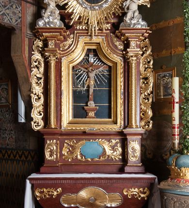 Zdjęcie nr 9: Ołtarz przyścienny, architektoniczny, jednokondygnacyjny, jednoosiowy, ze zwieńczeniem. Mensa w typie sarkofagowym z kamiennym portatylem, dekorowana w dolnej partii srebrnymi liśćmi akantu, a w górnej plakietą w kształcie leżącego prostokąta o zaokrąglonych narożach przenikającego owal; kształt wypełniają odchodzące od centralnego pola złote promienie. Ujmują je plakiety ornamentalne z kształtów nawiązujących do motywu rocaille’a; całość mensy groszkowana. Retabulum na cokole o wysuniętych bokach, ujęte parą kolumn o kapitelach akantowych i trzonach zdobionych żłobkowaniem od frontu, wspierających fragmenty belkowania z silnie wysuniętymi fryzem i gzymsem. Pola cokołu retabulum zdobione motywem wici akantowej ujmującej jasnoniebieski kartusz w kształcie leżącego owalu przenikającego się ze stojącym prostokątem. Oś środkową nastawy wypełnia pole w kształcie stojącego prostokąta o wklęsłych górnych narożach, ujęte profilowaną, częściowo złoconą ramą, z motywem liści laurowych. W środku wizerunek ukrzyżowanego Jezusa na tle lustra. Na zasuwie obraz św. Teresy od Dzieciątka Jezus. Kondygnację flankują ażurowe uszaki utworzone z bujnej, mięsistej wici z liści akantu. Zwieńczenie tworzy owalne przedstawienie św. Antoniego Padewskiego otoczone srebrnymi obłokami oraz glorią promienistą o promieniach różnej długości. Zwieńczenie flankuje para nagich aniołków posadowionych na srebrzonych chmurach, umieszczonych na fragmentach belkowania retabulum. 
Struktura polichromowana w kolorze brązowym. Partie detalu architektonicznego i ornamentyka złocone i srebrzone. 
