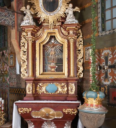 Zdjęcie nr 10: Ołtarz przyścienny, architektoniczny, jednokondygnacyjny, jednoosiowy, ze zwieńczeniem. Mensa w typie sarkofagowym z kamiennym portatylem, dekorowana w dolnej partii srebrnymi liśćmi akantu, a w górnej plakietą w kształcie leżącego prostokąta o zaokrąglonych narożach przenikającego owal; kształt wypełniają odchodzące od centralnego pola złote promienie. Ujmują je plakiety ornamentalne z kształtów nawiązujących do motywu rocaille’a; całość mensy groszkowana. Retabulum na cokole o wysuniętych bokach, ujęte parą kolumn o kapitelach akantowych i trzonach zdobionych żłobkowaniem od frontu, wspierających fragmenty belkowania z silnie wysuniętymi fryzem i gzymsem. Pola cokołu retabulum zdobione motywem wici akantowej ujmującej jasnoniebieski kartusz w kształcie leżącego owalu przenikającego się ze stojącym prostokątem. Oś środkową nastawy wypełnia pole w kształcie stojącego prostokąta o wklęsłych górnych narożach, ujęte profilowaną, częściowo złoconą ramą, z motywem liści laurowych. W środku wizerunek ukrzyżowanego Jezusa na tle lustra. Na zasuwie obraz św. Teresy od Dzieciątka Jezus. Kondygnację flankują ażurowe uszaki utworzone z bujnej, mięsistej wici z liści akantu. Zwieńczenie tworzy owalne przedstawienie św. Antoniego Padewskiego otoczone srebrnymi obłokami oraz glorią promienistą o promieniach różnej długości. Zwieńczenie flankuje para nagich aniołków posadowionych na srebrzonych chmurach, umieszczonych na fragmentach belkowania retabulum. 
Struktura polichromowana w kolorze brązowym. Partie detalu architektonicznego i ornamentyka złocone i srebrzone. 
