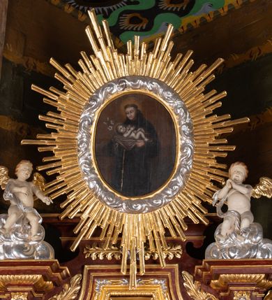 Zdjęcie nr 11: Ołtarz przyścienny, architektoniczny, jednokondygnacyjny, jednoosiowy, ze zwieńczeniem. Mensa w typie sarkofagowym z kamiennym portatylem, dekorowana w dolnej partii srebrnymi liśćmi akantu, a w górnej plakietą w kształcie leżącego prostokąta o zaokrąglonych narożach przenikającego owal; kształt wypełniają odchodzące od centralnego pola złote promienie. Ujmują je plakiety ornamentalne z kształtów nawiązujących do motywu rocaille’a; całość mensy groszkowana. Retabulum na cokole o wysuniętych bokach, ujęte parą kolumn o kapitelach akantowych i trzonach zdobionych żłobkowaniem od frontu, wspierających fragmenty belkowania z silnie wysuniętymi fryzem i gzymsem. Pola cokołu retabulum zdobione motywem wici akantowej ujmującej jasnoniebieski kartusz w kształcie leżącego owalu przenikającego się ze stojącym prostokątem. Oś środkową nastawy wypełnia pole w kształcie stojącego prostokąta o wklęsłych górnych narożach, ujęte profilowaną, częściowo złoconą ramą, z motywem liści laurowych. W środku wizerunek ukrzyżowanego Jezusa na tle lustra. Na zasuwie obraz św. Teresy od Dzieciątka Jezus. Kondygnację flankują ażurowe uszaki utworzone z bujnej, mięsistej wici z liści akantu. Zwieńczenie tworzy owalne przedstawienie św. Antoniego Padewskiego otoczone srebrnymi obłokami oraz glorią promienistą o promieniach różnej długości. Zwieńczenie flankuje para nagich aniołków posadowionych na srebrzonych chmurach, umieszczonych na fragmentach belkowania retabulum. 
Struktura polichromowana w kolorze brązowym. Partie detalu architektonicznego i ornamentyka złocone i srebrzone. 
