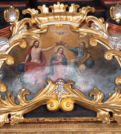 Zdjęcie nr 2: Obraz o nieregularnym kształcie zbliżonym do równoramiennego trójkąta o silnie zaokrąglonych wierzchołkach i wklęsłej podstawie, ujący złoconą i srebrzoną akantową ramą zwieńczoną koroną w typie otwartym. Ramę dekorują dodatkowo srebrzone winne grona. W centrum kompozycji, na tle obłoków ukazana klęcząca Maria z pochyloną głową i rękami skrzyżowanymi na piersi. Przedstawiona frontalnie jako młoda, długowłosa kobieta  z głową zwróconą w trzech czwartych w lewo. Twarz owalna, policzki delikatnie zaróżowione, wzrok skierowany w dół. Maria ubrana jest w jasnoróżową suknię z długimi rękawami oraz granatowy płaszcz luźno udrapowany wokół ramion postaci. Wokół głowy widoczny świetlisty nimb. Ponad głową, ukazani po bokach Marii Chrystus (z lewej) i Bóg Ojciec (z prawej) podtrzymują dużą, złotą koronę w typie otwartym. Nieco wyżej ukazana gołębica Ducha Świętego. Chrystus siedzący na obłoku przedstawiony frontalnie z twarzą skierowaną do Marii. Prawą rękę wspiera na niebieskim globie, lewą wyciąga, podtrzymując koronę nad głową swojej matki. Zbawiciel został przedstawiony jako młody mężczyzna o długich, brązowych włosach i takim samym zaroście. Jego tors z widocznymi śladami męki jest nagi, na ramiona i plecy na luźno narzucony czerwony płaszcz. Wokół jego głowy znajduje się świetlisty nimb. Przedstawiony z prawej strony obrazu Bóg Ojciec również siedzi w obłokach, zwracając się w kierunku Marii. Lewą rękę wspiera na niebieskim globie, trzymając w niej również złote berło. Prawą wyciągniętą w bok podtrzymuje koronę ponad głową Marii. Bóg Ojciec został przedstawiony jako starzec z siwymi włosami oraz długą siwą brodą ubrany w jasnoniebieską szatę z długimi rękawami oraz żółty płaszcz. W tle obrazu widoczne obłoki rozświetlone ciepłym, brązowo-żółtym światłem. Kolorystyka obrazu ciepła, intensywna.