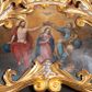 Zdjęcie nr 1: Obraz o nieregularnym kształcie zbliżonym do równoramiennego trójkąta o silnie zaokrąglonych wierzchołkach i wklęsłej podstawie, ujący złoconą i srebrzoną akantową ramą zwieńczoną koroną w typie otwartym. Ramę dekorują dodatkowo srebrzone winne grona. W centrum kompozycji, na tle obłoków ukazana klęcząca Maria z pochyloną głową i rękami skrzyżowanymi na piersi. Przedstawiona frontalnie jako młoda, długowłosa kobieta  z głową zwróconą w trzech czwartych w lewo. Twarz owalna, policzki delikatnie zaróżowione, wzrok skierowany w dół. Maria ubrana jest w jasnoróżową suknię z długimi rękawami oraz granatowy płaszcz luźno udrapowany wokół ramion postaci. Wokół głowy widoczny świetlisty nimb. Ponad głową, ukazani po bokach Marii Chrystus (z lewej) i Bóg Ojciec (z prawej) podtrzymują dużą, złotą koronę w typie otwartym. Nieco wyżej ukazana gołębica Ducha Świętego. Chrystus siedzący na obłoku przedstawiony frontalnie z twarzą skierowaną do Marii. Prawą rękę wspiera na niebieskim globie, lewą wyciąga, podtrzymując koronę nad głową swojej matki. Zbawiciel został przedstawiony jako młody mężczyzna o długich, brązowych włosach i takim samym zaroście. Jego tors z widocznymi śladami męki jest nagi, na ramiona i plecy na luźno narzucony czerwony płaszcz. Wokół jego głowy znajduje się świetlisty nimb. Przedstawiony z prawej strony obrazu Bóg Ojciec również siedzi w obłokach, zwracając się w kierunku Marii. Lewą rękę wspiera na niebieskim globie, trzymając w niej również złote berło. Prawą wyciągniętą w bok podtrzymuje koronę ponad głową Marii. Bóg Ojciec został przedstawiony jako starzec z siwymi włosami oraz długą siwą brodą ubrany w jasnoniebieską szatę z długimi rękawami oraz żółty płaszcz. W tle obrazu widoczne obłoki rozświetlone ciepłym, brązowo-żółtym światłem. Kolorystyka obrazu ciepła, intensywna.