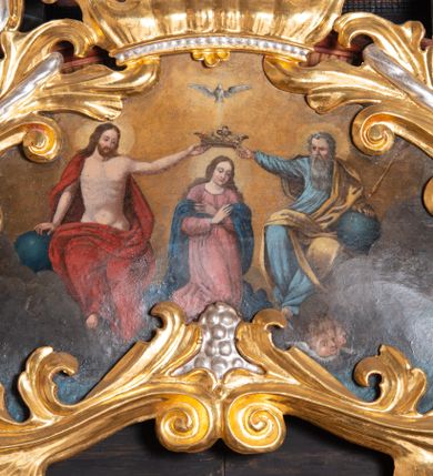 Zdjęcie nr 1: Obraz o nieregularnym kształcie zbliżonym do równoramiennego trójkąta o silnie zaokrąglonych wierzchołkach i wklęsłej podstawie, ujący złoconą i srebrzoną akantową ramą zwieńczoną koroną w typie otwartym. Ramę dekorują dodatkowo srebrzone winne grona. W centrum kompozycji, na tle obłoków ukazana klęcząca Maria z pochyloną głową i rękami skrzyżowanymi na piersi. Przedstawiona frontalnie jako młoda, długowłosa kobieta  z głową zwróconą w trzech czwartych w lewo. Twarz owalna, policzki delikatnie zaróżowione, wzrok skierowany w dół. Maria ubrana jest w jasnoróżową suknię z długimi rękawami oraz granatowy płaszcz luźno udrapowany wokół ramion postaci. Wokół głowy widoczny świetlisty nimb. Ponad głową, ukazani po bokach Marii Chrystus (z lewej) i Bóg Ojciec (z prawej) podtrzymują dużą, złotą koronę w typie otwartym. Nieco wyżej ukazana gołębica Ducha Świętego. Chrystus siedzący na obłoku przedstawiony frontalnie z twarzą skierowaną do Marii. Prawą rękę wspiera na niebieskim globie, lewą wyciąga, podtrzymując koronę nad głową swojej matki. Zbawiciel został przedstawiony jako młody mężczyzna o długich, brązowych włosach i takim samym zaroście. Jego tors z widocznymi śladami męki jest nagi, na ramiona i plecy na luźno narzucony czerwony płaszcz. Wokół jego głowy znajduje się świetlisty nimb. Przedstawiony z prawej strony obrazu Bóg Ojciec również siedzi w obłokach, zwracając się w kierunku Marii. Lewą rękę wspiera na niebieskim globie, trzymając w niej również złote berło. Prawą wyciągniętą w bok podtrzymuje koronę ponad głową Marii. Bóg Ojciec został przedstawiony jako starzec z siwymi włosami oraz długą siwą brodą ubrany w jasnoniebieską szatę z długimi rękawami oraz żółty płaszcz. W tle obrazu widoczne obłoki rozświetlone ciepłym, brązowo-żółtym światłem. Kolorystyka obrazu ciepła, intensywna.