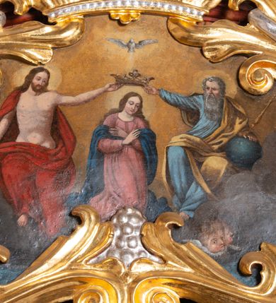 Zdjęcie nr 4: Obraz o nieregularnym kształcie zbliżonym do równoramiennego trójkąta o silnie zaokrąglonych wierzchołkach i wklęsłej podstawie, ujący złoconą i srebrzoną akantową ramą zwieńczoną koroną w typie otwartym. Ramę dekorują dodatkowo srebrzone winne grona. W centrum kompozycji, na tle obłoków ukazana klęcząca Maria z pochyloną głową i rękami skrzyżowanymi na piersi. Przedstawiona frontalnie jako młoda, długowłosa kobieta  z głową zwróconą w trzech czwartych w lewo. Twarz owalna, policzki delikatnie zaróżowione, wzrok skierowany w dół. Maria ubrana jest w jasnoróżową suknię z długimi rękawami oraz granatowy płaszcz luźno udrapowany wokół ramion postaci. Wokół głowy widoczny świetlisty nimb. Ponad głową, ukazani po bokach Marii Chrystus (z lewej) i Bóg Ojciec (z prawej) podtrzymują dużą, złotą koronę w typie otwartym. Nieco wyżej ukazana gołębica Ducha Świętego. Chrystus siedzący na obłoku przedstawiony frontalnie z twarzą skierowaną do Marii. Prawą rękę wspiera na niebieskim globie, lewą wyciąga, podtrzymując koronę nad głową swojej matki. Zbawiciel został przedstawiony jako młody mężczyzna o długich, brązowych włosach i takim samym zaroście. Jego tors z widocznymi śladami męki jest nagi, na ramiona i plecy na luźno narzucony czerwony płaszcz. Wokół jego głowy znajduje się świetlisty nimb. Przedstawiony z prawej strony obrazu Bóg Ojciec również siedzi w obłokach, zwracając się w kierunku Marii. Lewą rękę wspiera na niebieskim globie, trzymając w niej również złote berło. Prawą wyciągniętą w bok podtrzymuje koronę ponad głową Marii. Bóg Ojciec został przedstawiony jako starzec z siwymi włosami oraz długą siwą brodą ubrany w jasnoniebieską szatę z długimi rękawami oraz żółty płaszcz. W tle obrazu widoczne obłoki rozświetlone ciepłym, brązowo-żółtym światłem. Kolorystyka obrazu ciepła, intensywna.
