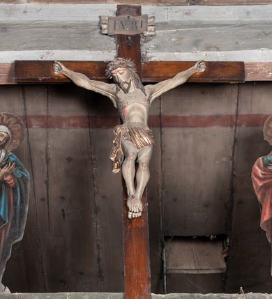 Zdjęcie nr 3: Chrystus rozpięty na krzyżu z szeroko rozłożonymi rękami, w lekkim zwisie ciała, z lekko ugiętymi kolanami i stopami założonymi prawa na lewą, przybitymi jednym gwoździem. Głowa pochylona na prawy bark. Twarz pociągła z długim nosem i wydatnymi kośćmi policzkowymi, okolona długimi ciemnymi włosami i zarostem. Ciało muskularne o ciemnej karnacji z zaznaczonymi śladami męki. Wokół bioder złocone perizonium podtrzymywane sznurem, ze zwisem draperii wzdłuż prawego uda. Matka Boska Bolesna ukazana w całej postaci, w ujęciu w trzech czwartych, zwrócona w lewo w kierunku krzyża; stoi w kontrapoście z głową przechyloną w lewo. Ręce ugięte w łokciach, dłonie skrzyżowane w przegubach ułożone na wysokości piersi. Ubrana w długą fałdzistą czerwoną suknię i narzucony na ramiona ciemnoniebieski płaszcz. Na głowie i ramionach biała chusta; wokół głowy nimb. Św. Jan Ewangelista przedstawiony w całej postaci, w ujęciu w trzech czwartych, zwrócony w prawo w kierunku krzyża.  Twarz o młodzieńczych rysach (bez zarostu), widoczna z profilu, zwrócona ku górze, ku Chrystusowi. Ręce ugięte w łokciach, z dłońmi złożonymi na piersiach na różnej wysokości. Ubrany w długą ciemnoniebieską suknię i czerwony płaszcz. Wokół głowy nimb.   