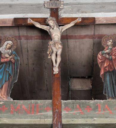 Zdjęcie nr 1: Chrystus rozpięty na krzyżu z szeroko rozłożonymi rękami, w lekkim zwisie ciała, z lekko ugiętymi kolanami i stopami założonymi prawa na lewą, przybitymi jednym gwoździem. Głowa pochylona na prawy bark. Twarz pociągła z długim nosem i wydatnymi kośćmi policzkowymi, okolona długimi ciemnymi włosami i zarostem. Ciało muskularne o ciemnej karnacji z zaznaczonymi śladami męki. Wokół bioder złocone perizonium podtrzymywane sznurem, ze zwisem draperii wzdłuż prawego uda. Matka Boska Bolesna ukazana w całej postaci, w ujęciu w trzech czwartych, zwrócona w lewo w kierunku krzyża; stoi w kontrapoście z głową przechyloną w lewo. Ręce ugięte w łokciach, dłonie skrzyżowane w przegubach ułożone na wysokości piersi. Ubrana w długą fałdzistą czerwoną suknię i narzucony na ramiona ciemnoniebieski płaszcz. Na głowie i ramionach biała chusta; wokół głowy nimb. Św. Jan Ewangelista przedstawiony w całej postaci, w ujęciu w trzech czwartych, zwrócony w prawo w kierunku krzyża.  Twarz o młodzieńczych rysach (bez zarostu), widoczna z profilu, zwrócona ku górze, ku Chrystusowi. Ręce ugięte w łokciach, z dłońmi złożonymi na piersiach na różnej wysokości. Ubrany w długą ciemnoniebieską suknię i czerwony płaszcz. Wokół głowy nimb.   