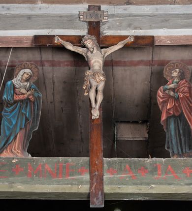 Zdjęcie nr 2: Chrystus rozpięty na krzyżu z szeroko rozłożonymi rękami, w lekkim zwisie ciała, z lekko ugiętymi kolanami i stopami założonymi prawa na lewą, przybitymi jednym gwoździem. Głowa pochylona na prawy bark. Twarz pociągła z długim nosem i wydatnymi kośćmi policzkowymi, okolona długimi ciemnymi włosami i zarostem. Ciało muskularne o ciemnej karnacji z zaznaczonymi śladami męki. Wokół bioder złocone perizonium podtrzymywane sznurem, ze zwisem draperii wzdłuż prawego uda. Matka Boska Bolesna ukazana w całej postaci, w ujęciu w trzech czwartych, zwrócona w lewo w kierunku krzyża; stoi w kontrapoście z głową przechyloną w lewo. Ręce ugięte w łokciach, dłonie skrzyżowane w przegubach ułożone na wysokości piersi. Ubrana w długą fałdzistą czerwoną suknię i narzucony na ramiona ciemnoniebieski płaszcz. Na głowie i ramionach biała chusta; wokół głowy nimb. Św. Jan Ewangelista przedstawiony w całej postaci, w ujęciu w trzech czwartych, zwrócony w prawo w kierunku krzyża.  Twarz o młodzieńczych rysach (bez zarostu), widoczna z profilu, zwrócona ku górze, ku Chrystusowi. Ręce ugięte w łokciach, z dłońmi złożonymi na piersiach na różnej wysokości. Ubrany w długą ciemnoniebieską suknię i czerwony płaszcz. Wokół głowy nimb.   