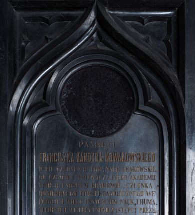 Zdjęcie nr 1: Epitafium w formie stojącego prostokąta,  ujęte szerokim obramieniem, z głęboką płyciną z motywem oślego łuku z wpisanym w niego maswerkiem oraz z maswerkami w nadłuczach. Nad górną krawędzią ujęty dwiema ulistnionymi wolutkami krzyżyk łaciński o fazowanych krawędziach. W łuku okrągła płycina z polem na portret, poniżej inskrypcja „PAMIĘCI / FRANCISZKA KANDYDA NOWAKOWSKIEGO / D(OKTO)RA FIL(OZOFII) CZŁONKA B(IERNEGO) TOW(ARZYSTWA) NAUK(OWEGO) KRAKOWSKIE/GO CZŁONKA NADZWYCZAJNEGO AKADEMII / UMIEJĘTNOŚCI W KRAKOWIE_CZŁONKA / HONOROWEGO TOW(ARZYSTWA) PEDAGOGICZNEGO WE / LWOWIE I WIELU INNYCH TOW(ARZYSTW) NAUK(OWYCH) I HUMA/NITARNYCH. WIELOLETNEGO ZASTĘPCY PREZE/SA RADY SZKOLNEJ OKRĘGU WADOWICKIEGO ZA/SŁUŻONEGO AUTORA NA POLU PEDAGOGII, I GOR/LIWEGO MIŁOŚNIKA OJCZYSTEJ MOWY. / „CHODZIŁ W OCZACH TWOICH PANIE W PRAWDZIE I SPRAWIEDLIWOŚCI / PRAWEM SERCEM BYŁ Z TOBĄ, A TY ZACHOWAŁEŚ MU MIŁOSIERDZIE TWOJE&quot;. / Z KS(IĘGI) III KRÓL(EWSKIEJ) ROZ(DZIAŁ) 3. / *3 X 1813 W POZNANIU † 19 XI 1881 W SUCHY. / NAJZACNIEJSZEMU MĘŻOWI ŻONA / PROSI POBOŻNYCH  O MODLITWĘ.”
