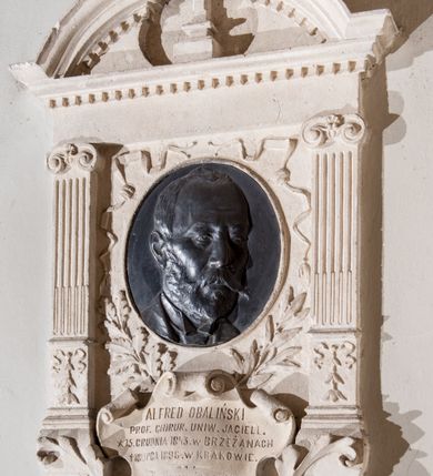 Zdjęcie nr 2: Epitafium w formie aediculi, flankowane przez kanelowane pilastry z jońskimi kapitelami. Powyżej pełne belkowanie z wysokim fryzem, gzymsem ozdobionym kostkowaniem i przerwanym przyczółkiem w kształcie luku nadwieszonego, z krzyżem łacińskim pośrodku. W polu medalion z płaskorzeźbionym portretem zmarłego, otoczony tasiemkami i liśćmi laurowymi oraz dębowymi. Poniżej, w kartuszu, inskrypcja: &quot;ALFRED OBALIŃSKI / PROF(ESOR) CHIRUR(GII) UNIW(ERSYTETU) JAGIELL(OŃSKIEGO) / * 15. GRUDNIA 1843. W BRZEŻANACH / † 18 LIPCA 1898 W KRAKOWIE. / Ku wiecznej pamięci / kamień ten położyli koledzy / Tow(arzystwa) Lek(arskiego) Krakowskiego / i Prof(esorowie) Uniwersytetu&quot;.