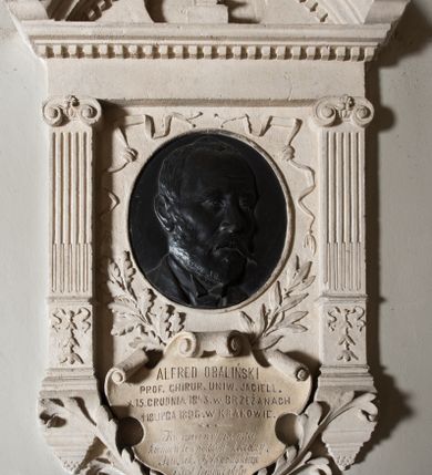 Zdjęcie nr 1: Epitafium w formie aediculi, flankowane przez kanelowane pilastry z jońskimi kapitelami. Powyżej pełne belkowanie z wysokim fryzem, gzymsem ozdobionym kostkowaniem i przerwanym przyczółkiem w kształcie luku nadwieszonego, z krzyżem łacińskim pośrodku. W polu medalion z płaskorzeźbionym portretem zmarłego, otoczony tasiemkami i liśćmi laurowymi oraz dębowymi. Poniżej, w kartuszu, inskrypcja: "ALFRED OBALIŃSKI / PROF(ESOR) CHIRUR(GII) UNIW(ERSYTETU) JAGIELL(OŃSKIEGO) / * 15. GRUDNIA 1843. W BRZEŻANACH / † 18 LIPCA 1898 W KRAKOWIE. / Ku wiecznej pamięci / kamień ten położyli koledzy / Tow(arzystwa) Lek(arskiego) Krakowskiego / i Prof(esorowie) Uniwersytetu".