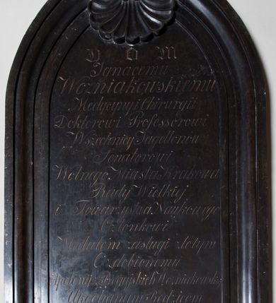 Zdjęcie nr 2: Epitafium w kształcie stojącego prostokąta, zamkniętego dwiema, półkolistymi, stykającymi się wolutami, połączonymi zwróconą ku dołowi muszlą  i zwieńczonymi krzyżem o ramionach zakończonych trójlistnie. W polu inskrypcja „D[EO] O[PTIMO] M[AXIMO] / Ignacemu / Wozniakowskiemu / Medycyny i Chirurgii / Doktorowi Professorowi / Wszechnicy Jagellonów / Senatorowi / Wolnego Miasta Krakowa / Rady Wielkiej / i Towarzystwa Naukowego / Członkowi / Medalem zasługi złotym / Ozdobionemiu | Apolonij z Rogujskich Wozniakowskiej / Ukochanym Rodzicom / Ignacy i Dawid / wdzięczni Synowie / r(oku) Pań(skiego) 1849.”
