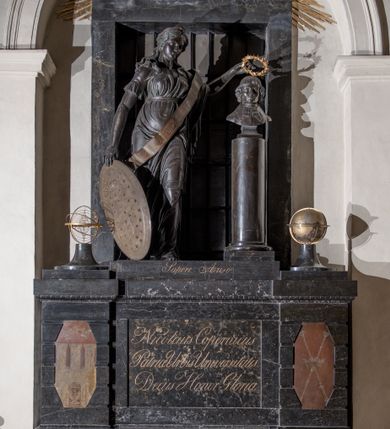 Zdjęcie nr 2: Pomnik przyścienny składa się z wysokiego cokołu, na którym stoi kobieta nakładająca wieniec laurowy na popiersie Mikołaja Kopernika osadzone na kolumnie. Cokół ujęty jest dwoma boniowanymi ryzalitami, z ośmiobocznymi tarczami z herbem Krakowa i Uniwersytetu Krakowskiego. Pośrodku cokołu wyryto inskrypcję: &quot;Nicolaus Copernicus / Patriae Urbis Universisitatis / Decus Honor Gloria&quot;. Po bokach na cokole ustawiono na niskich zwężających się, okrągłych w planie cokolikach glob ziemski i sferę planet. Pomiędzy nimi niska płyta z inskrypcją &quot;Sapere Auso&quot;. Nad niej stoi kobieta, personifikacja Uranii (?) ukazana w kontrapoście, w szatach ściśle przylegających do ciała i z szarfą przepasaną przez lewe ramię ze znakami zodiaku.  Jej twarz jest owalna, o zaostrzonej brodzie, ostrym nosie i drobnych ustach, otwartych, wydanych oczach, z włosami gęstymi, długimi, falującymi lokami opadającymi na ramiona. Głowa zwieńczona diademem z gwiazdą. Prawą ręką trzyma Tarczę Sobieskiego (z herbem Janina i astrologicznym Bykiem wśród gwiazd) opartą o podłoże, a w lewej wieniec z lauru nad głową Kopernika. Popiersie astronoma jest osadzone na wysokim cokole w kształcie gładkiej kolumny, ukazane frontalnie. Twarz modelowana sumarycznie, z zaznaczonymi zmarszczkami, prostym nosem i kręconymi włosami sięgającymi podbródka. Grupa rzeźbiarska ukazana na tle prostokątnego, zamurowanego przejścia, z kratą. Nad nim złocona gloria z kłębiastymi chmurami, między którymi napis „STA SOL NE MOVERE”. 