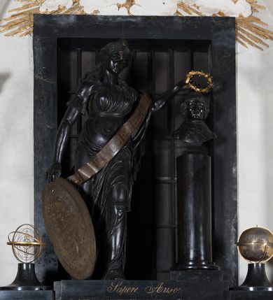 Zdjęcie nr 4: Pomnik przyścienny składa się z wysokiego cokołu, na którym stoi kobieta nakładająca wieniec laurowy na popiersie Mikołaja Kopernika osadzone na kolumnie. Cokół ujęty jest dwoma boniowanymi ryzalitami, z ośmiobocznymi tarczami z herbem Krakowa i Uniwersytetu Krakowskiego. Pośrodku cokołu wyryto inskrypcję: "Nicolaus Copernicus / Patriae Urbis Universisitatis / Decus Honor Gloria". Po bokach na cokole ustawiono na niskich zwężających się, okrągłych w planie cokolikach glob ziemski i sferę planet. Pomiędzy nimi niska płyta z inskrypcją "Sapere Auso". Nad niej stoi kobieta, personifikacja Uranii (?) ukazana w kontrapoście, w szatach ściśle przylegających do ciała i z szarfą przepasaną przez lewe ramię ze znakami zodiaku.  Jej twarz jest owalna, o zaostrzonej brodzie, ostrym nosie i drobnych ustach, otwartych, wydanych oczach, z włosami gęstymi, długimi, falującymi lokami opadającymi na ramiona. Głowa zwieńczona diademem z gwiazdą. Prawą ręką trzyma Tarczę Sobieskiego (z herbem Janina i astrologicznym Bykiem wśród gwiazd) opartą o podłoże, a w lewej wieniec z lauru nad głową Kopernika. Popiersie astronoma jest osadzone na wysokim cokole w kształcie gładkiej kolumny, ukazane frontalnie. Twarz modelowana sumarycznie, z zaznaczonymi zmarszczkami, prostym nosem i kręconymi włosami sięgającymi podbródka. Grupa rzeźbiarska ukazana na tle prostokątnego, zamurowanego przejścia, z kratą. Nad nim złocona gloria z kłębiastymi chmurami, między którymi napis „STA SOL NE MOVERE”. 