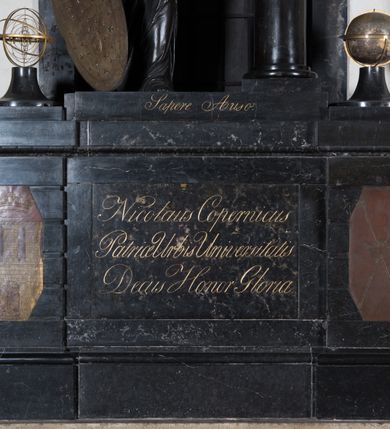 Zdjęcie nr 5: Pomnik przyścienny składa się z wysokiego cokołu, na którym stoi kobieta nakładająca wieniec laurowy na popiersie Mikołaja Kopernika osadzone na kolumnie. Cokół ujęty jest dwoma boniowanymi ryzalitami, z ośmiobocznymi tarczami z herbem Krakowa i Uniwersytetu Krakowskiego. Pośrodku cokołu wyryto inskrypcję: &quot;Nicolaus Copernicus / Patriae Urbis Universisitatis / Decus Honor Gloria&quot;. Po bokach na cokole ustawiono na niskich zwężających się, okrągłych w planie cokolikach glob ziemski i sferę planet. Pomiędzy nimi niska płyta z inskrypcją &quot;Sapere Auso&quot;. Nad niej stoi kobieta, personifikacja Uranii (?) ukazana w kontrapoście, w szatach ściśle przylegających do ciała i z szarfą przepasaną przez lewe ramię ze znakami zodiaku.  Jej twarz jest owalna, o zaostrzonej brodzie, ostrym nosie i drobnych ustach, otwartych, wydanych oczach, z włosami gęstymi, długimi, falującymi lokami opadającymi na ramiona. Głowa zwieńczona diademem z gwiazdą. Prawą ręką trzyma Tarczę Sobieskiego (z herbem Janina i astrologicznym Bykiem wśród gwiazd) opartą o podłoże, a w lewej wieniec z lauru nad głową Kopernika. Popiersie astronoma jest osadzone na wysokim cokole w kształcie gładkiej kolumny, ukazane frontalnie. Twarz modelowana sumarycznie, z zaznaczonymi zmarszczkami, prostym nosem i kręconymi włosami sięgającymi podbródka. Grupa rzeźbiarska ukazana na tle prostokątnego, zamurowanego przejścia, z kratą. Nad nim złocona gloria z kłębiastymi chmurami, między którymi napis „STA SOL NE MOVERE”. 